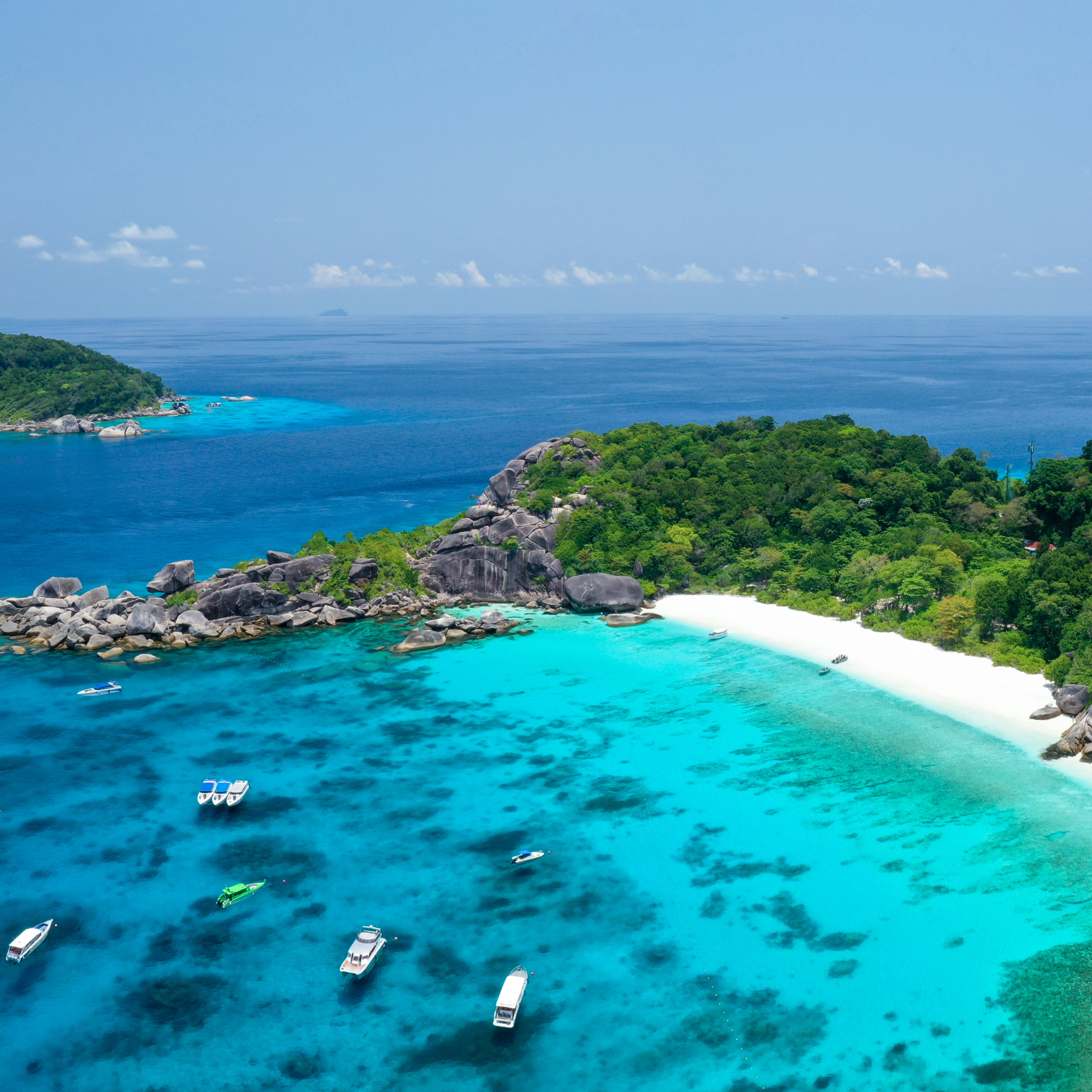 Vista aérea de un paraíso tropical con aguas turquesas, una playa de arena blanca y vegetación exuberante, varias embarcaciones en el mar y un cielo despejado con pocas nubes.