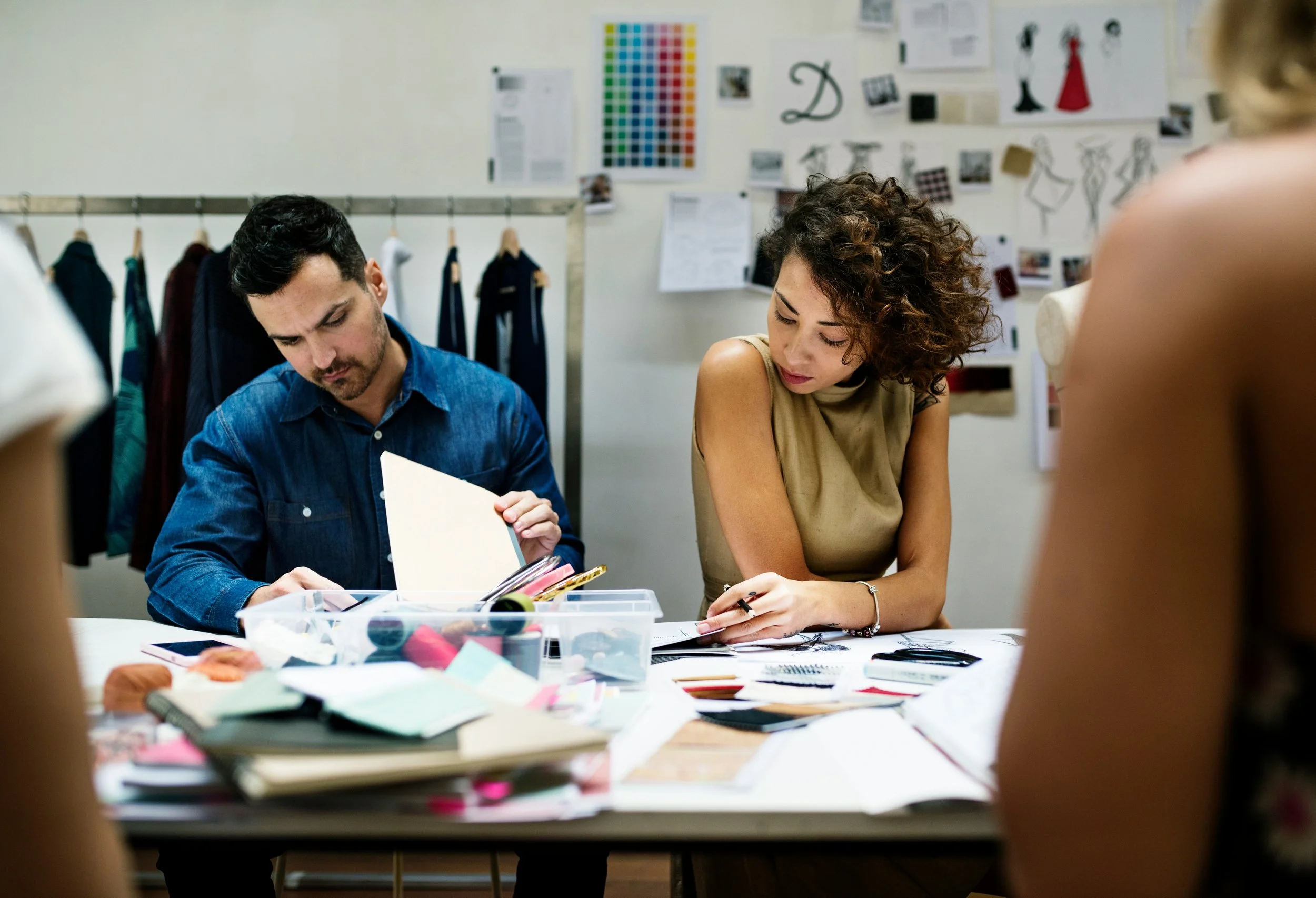 A man and a woman sitting at a table cluttered with papers and art supplies, working on a project in a creative studio.