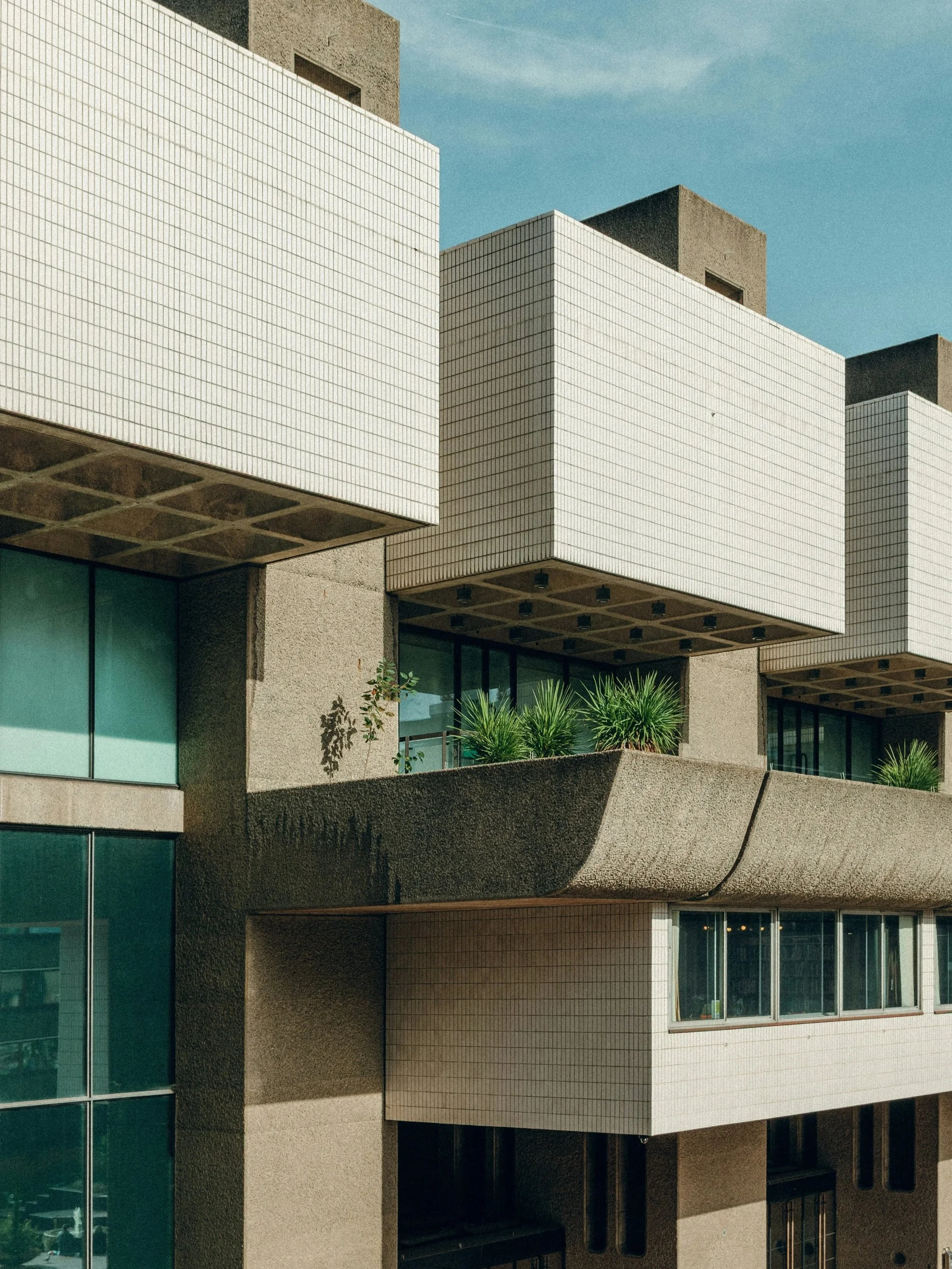 Close-up of a modern building with white tiled and concrete exterior, featuring balconies with green plants and large glass windows, under a blue sky.