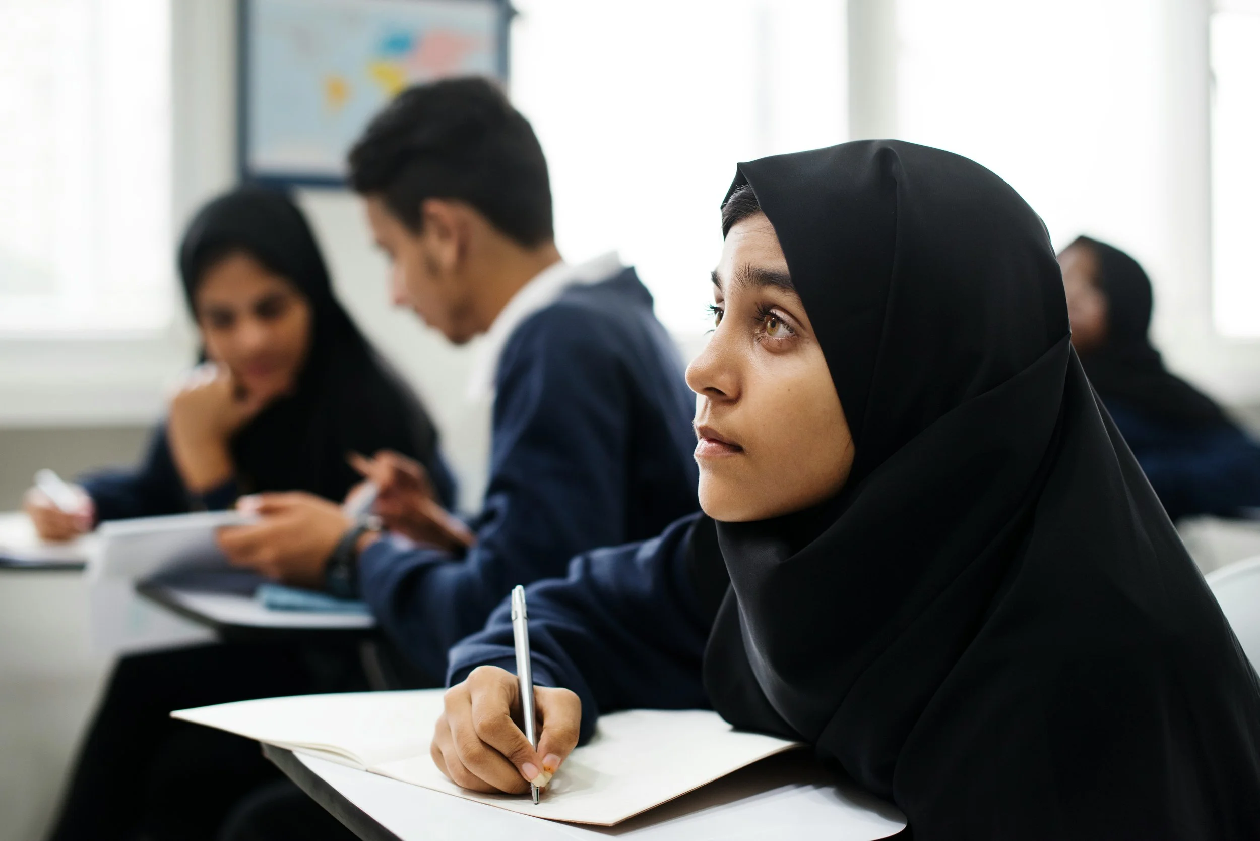 A young woman wearing a black hijab taking notes during a classroom lecture with other students in the background.