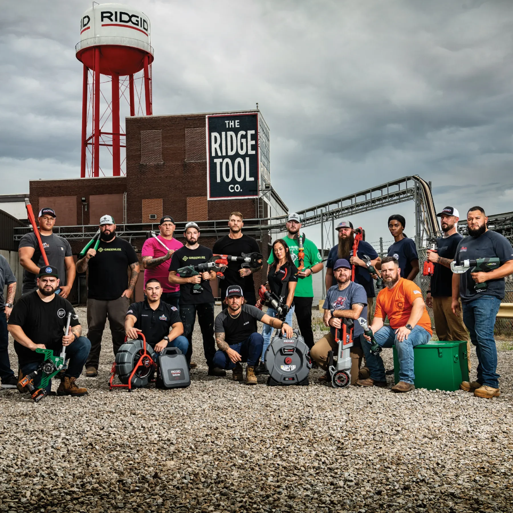 Group of people in front of a large brick building with a sign that reads "The Ridge Tool Co." and a water tower marked "RIDGID" on a cloudy day. They are holding various power tools and equipment.