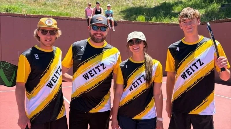 Four young tennis players standing on a tennis court, wearing black, yellow, and white jerseys with 'WEITZ' on them, smiling at the camera, with spectators sitting on a hill in the background.