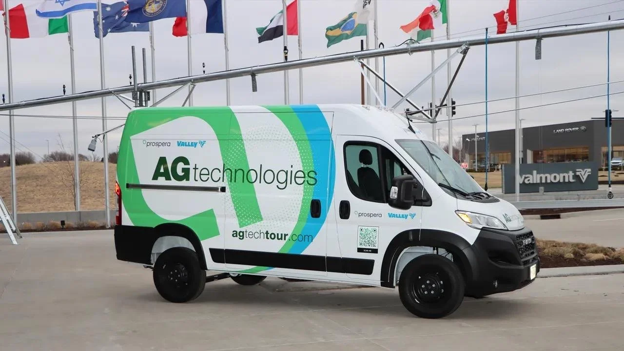 A white van with AG Technologies branding and website URL parked outside at a dealership, with flags flying overhead and a Land Rover dealership in the background.