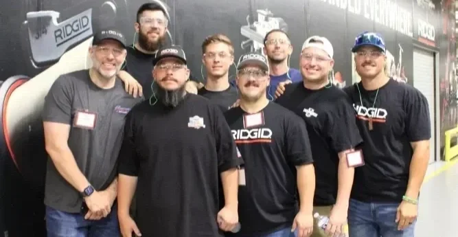 Group of eight men posing together at an event, some wearing branded T-shirts and hats, standing in front of a promotional backdrop.