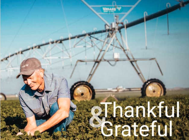 Farmer tending to crops in a field with a large irrigation sprinkler system in the background. The sky is clear and blue, and the scene conveys a sense of gratitude and hard work.