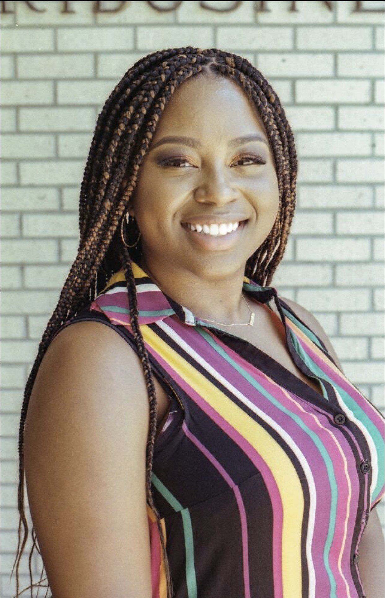 A woman with long braided hair smiling at the camera standing in front of a light-colored brick wall.