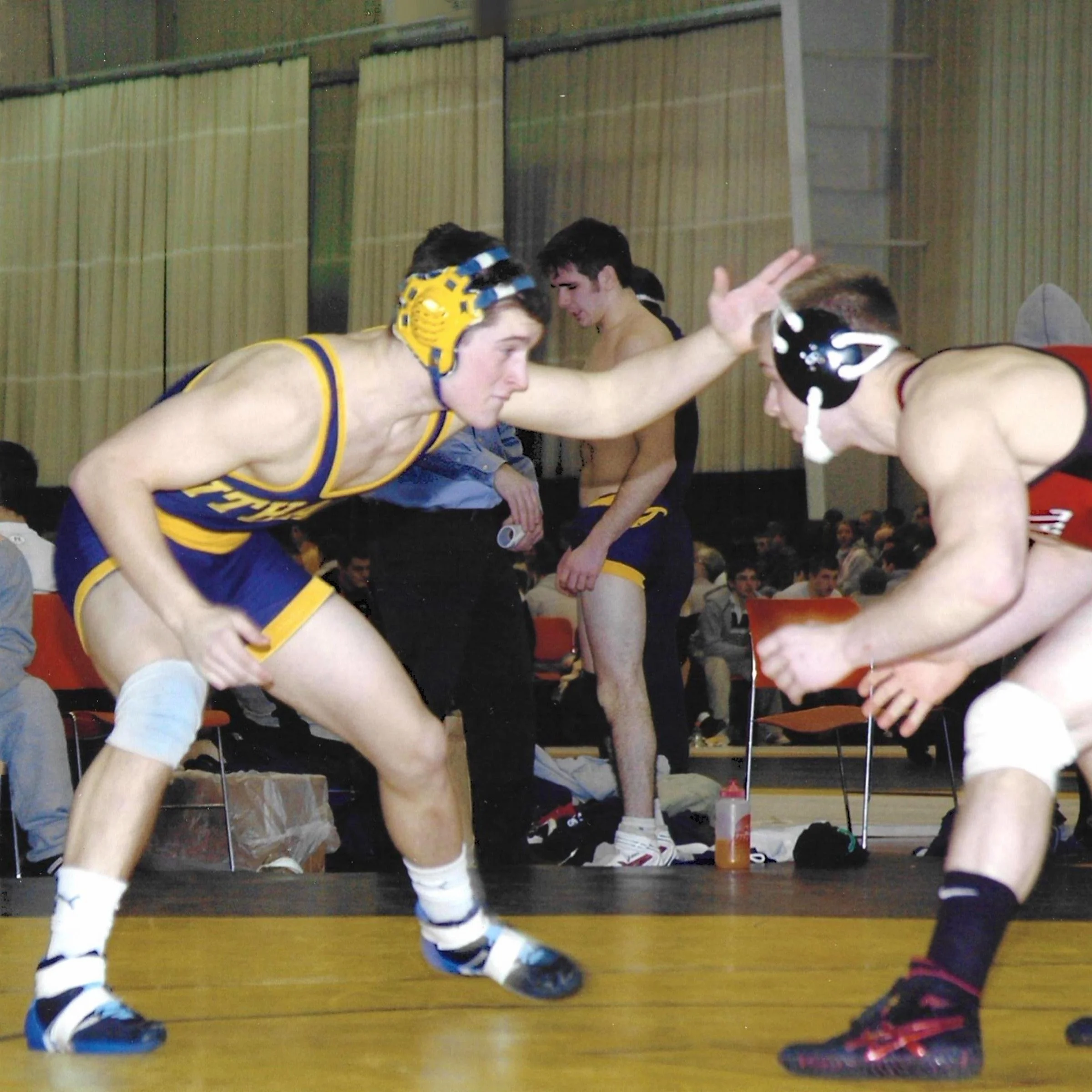 John Dale wrestling in a college match, in a blue and gold singlet and headgear, squaring up against an opponent in a neutral stance.
