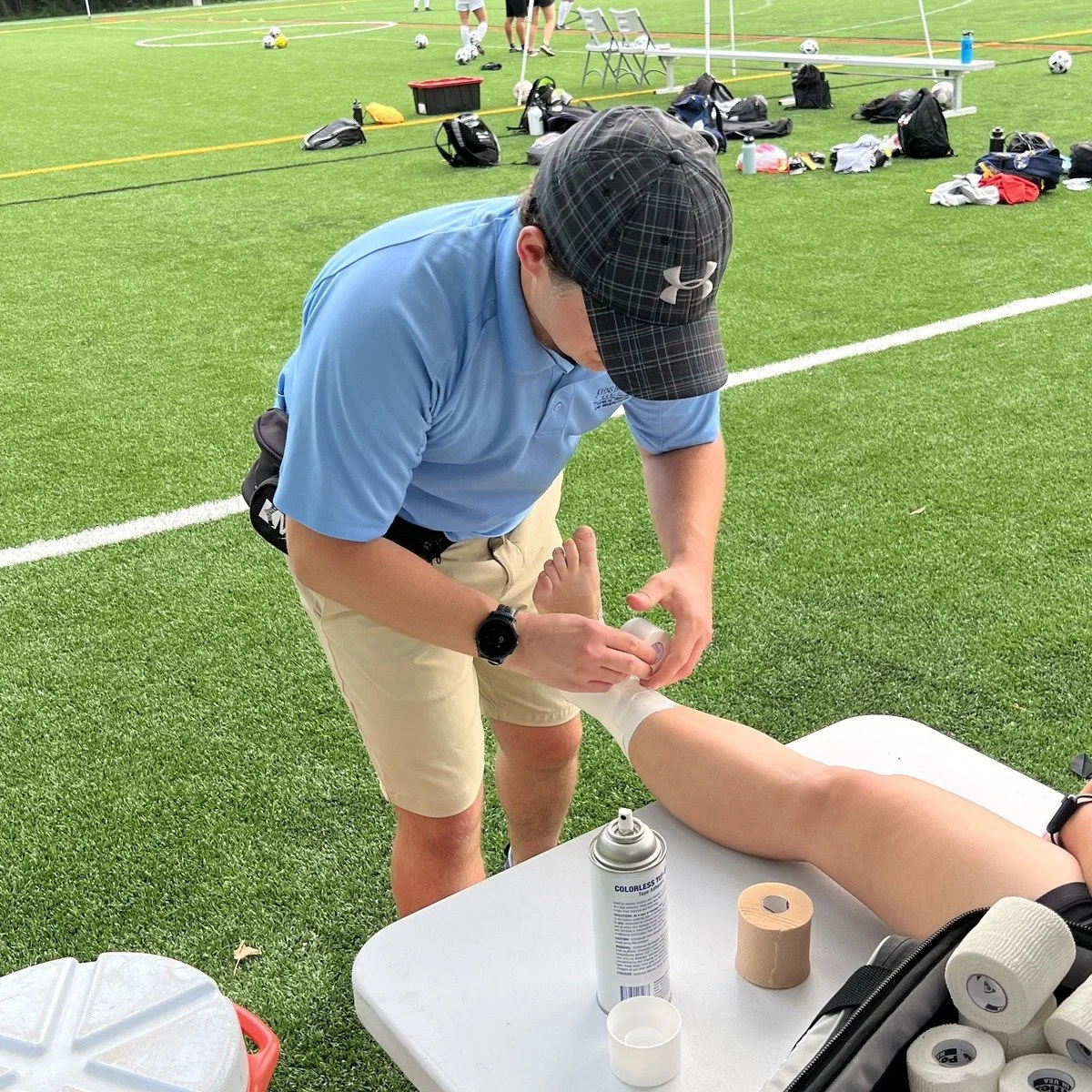 John Dale taping an athlete's ankle on the sideline of a turf field, working as a certified athletic trainer.
