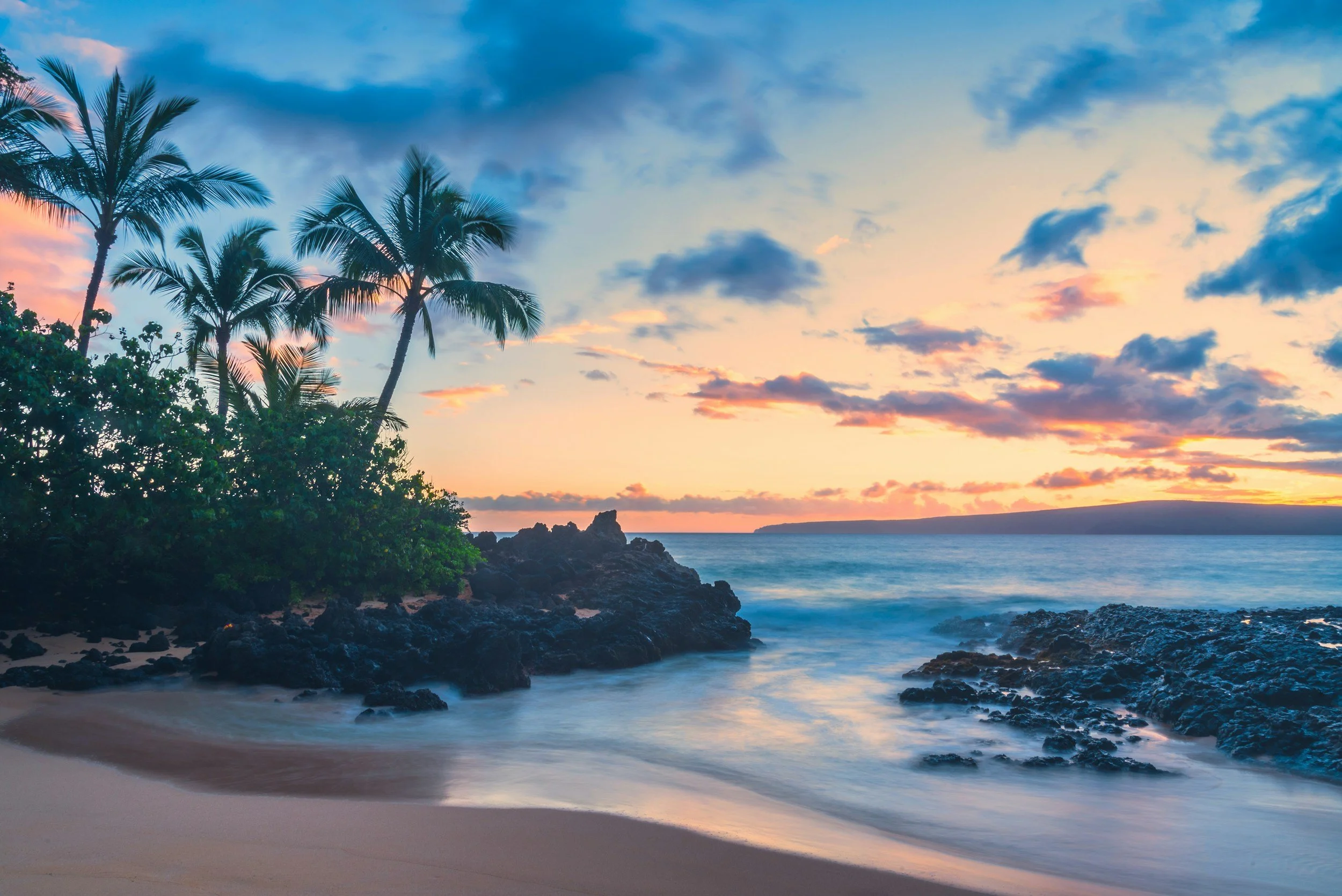 Palm trees frame a peaceful tropical beach at sunset with calm ocean water and pink and orange clouds.