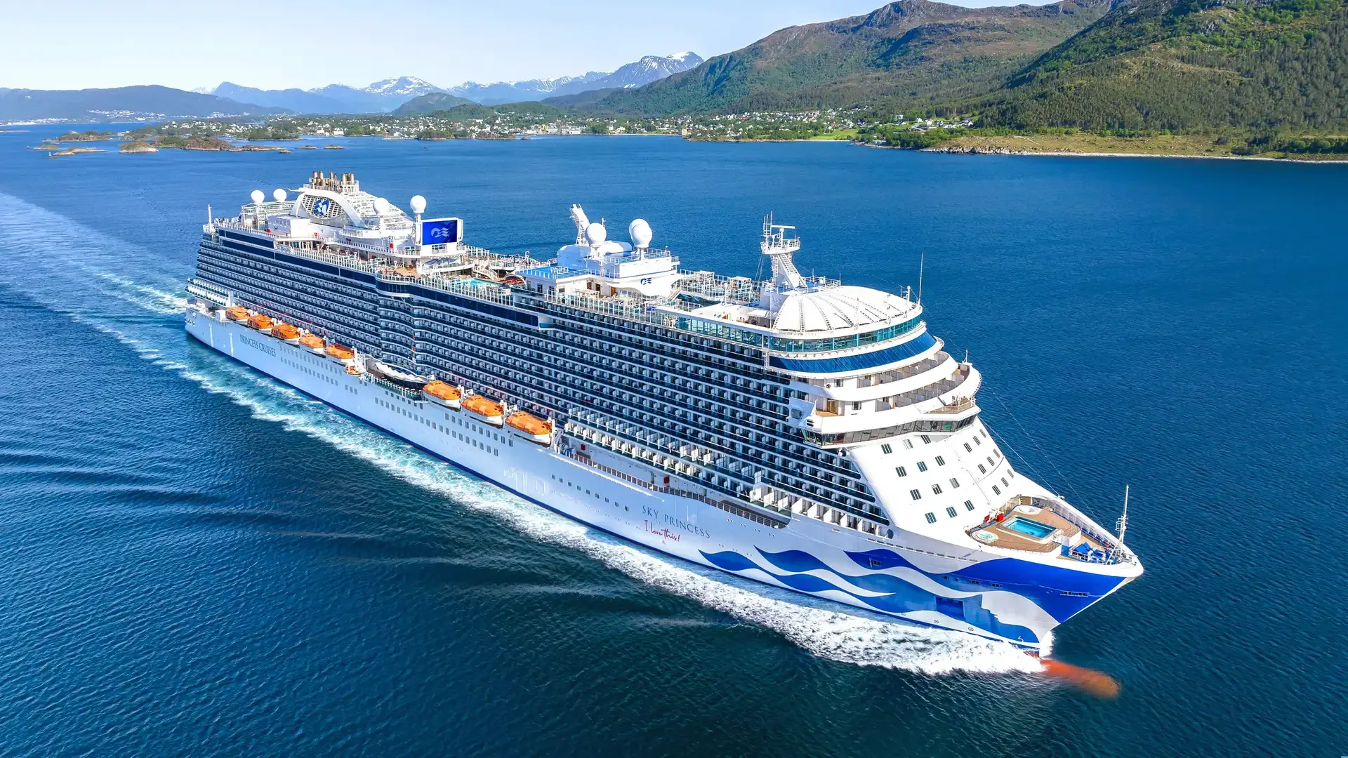 Sky Princess cruise ship sailing through calm blue water beside green mountains and snow-capped peaks in Alaska.