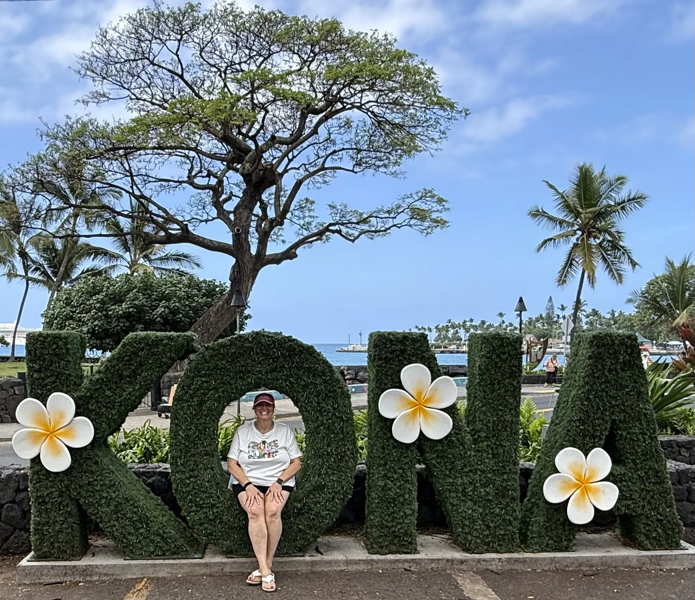 Janell Hammersberg seated in front of a tropical greenery wall decorated with white plumeria flowers beside the water.