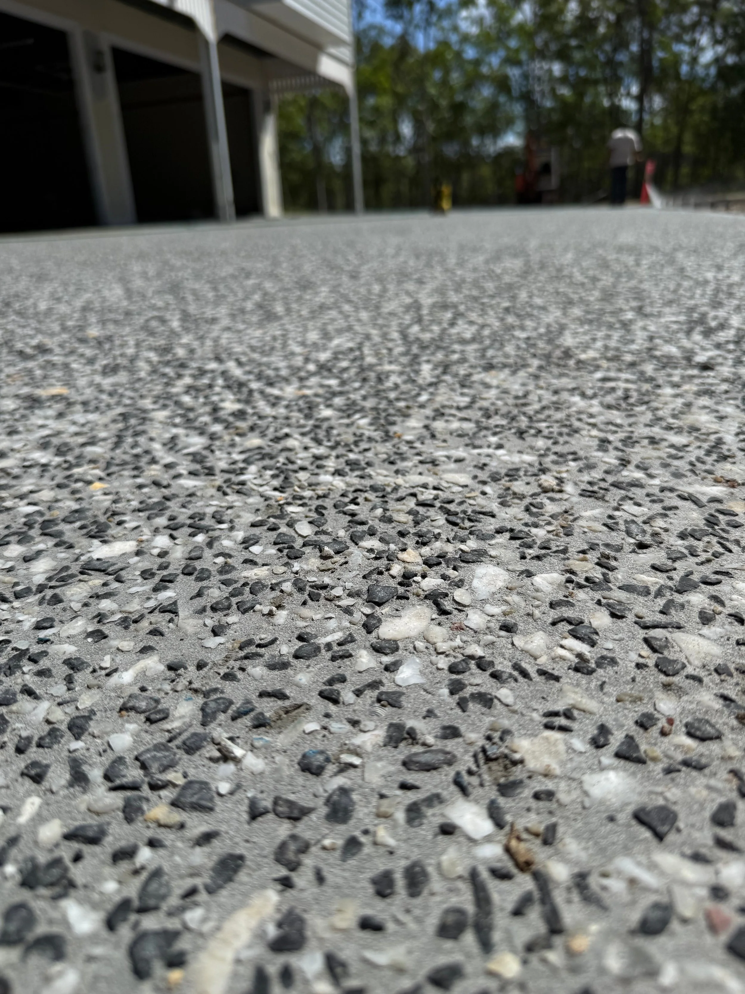 Close-up view of a gravelly concrete sidewalk surface with a building on the left and trees in the background.
