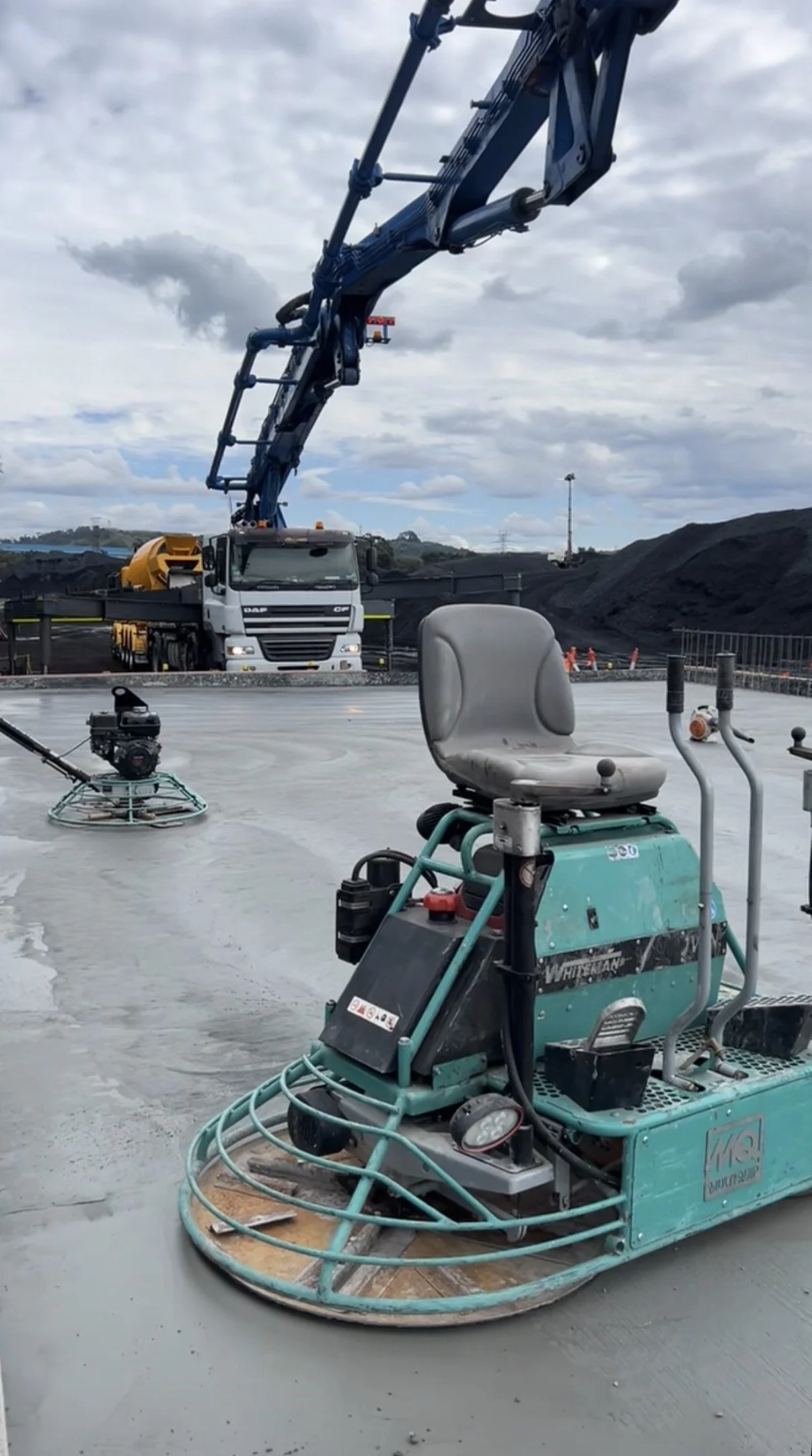 Construction site with a concrete polishing machine in the foreground and a crane pouring concrete in the background. Overcast sky with gray clouds.