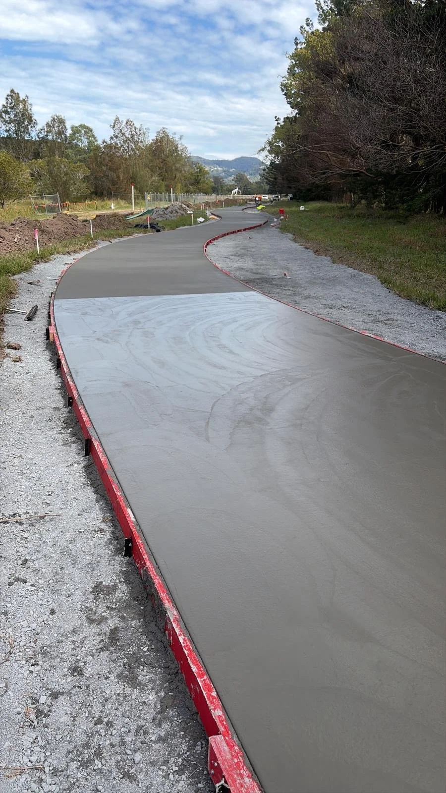 A newly paved winding road under construction, with barriers on the side and construction equipment in the distance, surrounded by trees and hills under a partly cloudy sky.