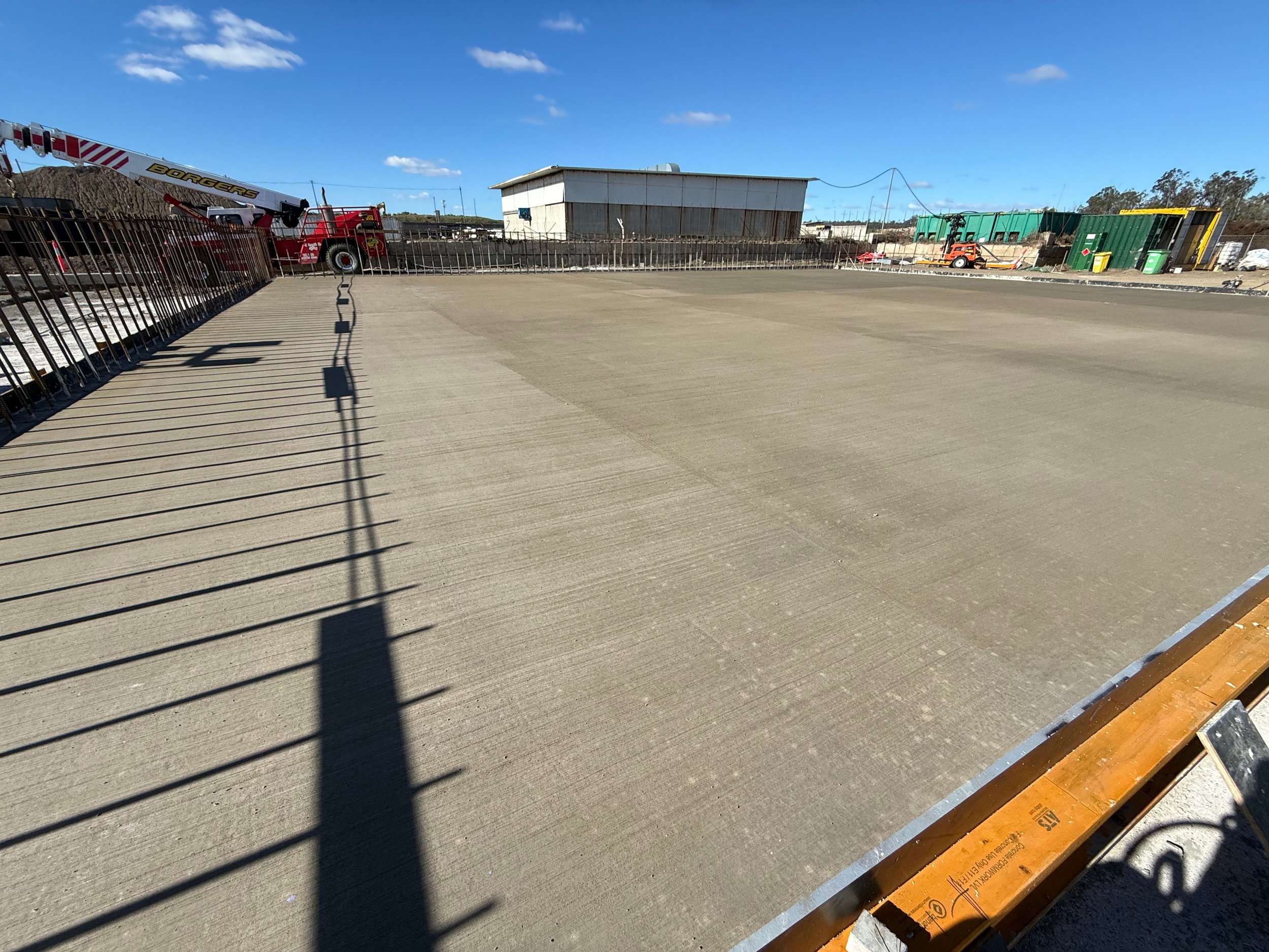 Construction site with a concrete floor, safety barriers, and scattered equipment, with a blue sky and a small building in the background.