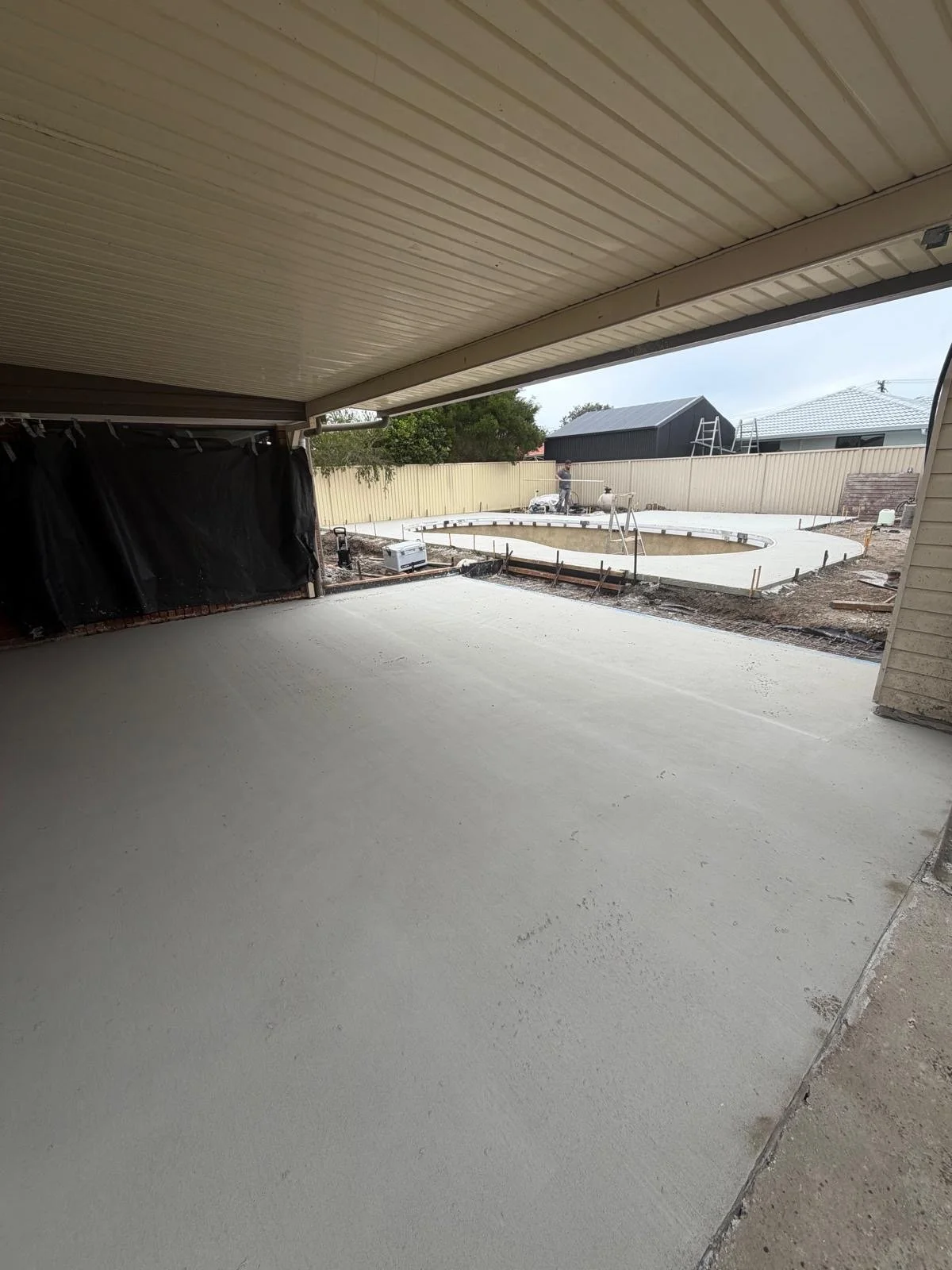 View from a covered patio looking out to a backyard garden with a swimming pool under construction. The patio floor appears freshly poured and smooth. The backyard is enclosed by a beige fence, and there is a black shed with a ladder on its roof in t