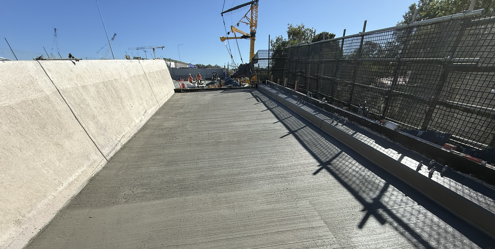 Construction site of a bridge or similar structure with a concrete surface, workers in safety gear, a crane, and safety fencing along the edges under a clear blue sky.