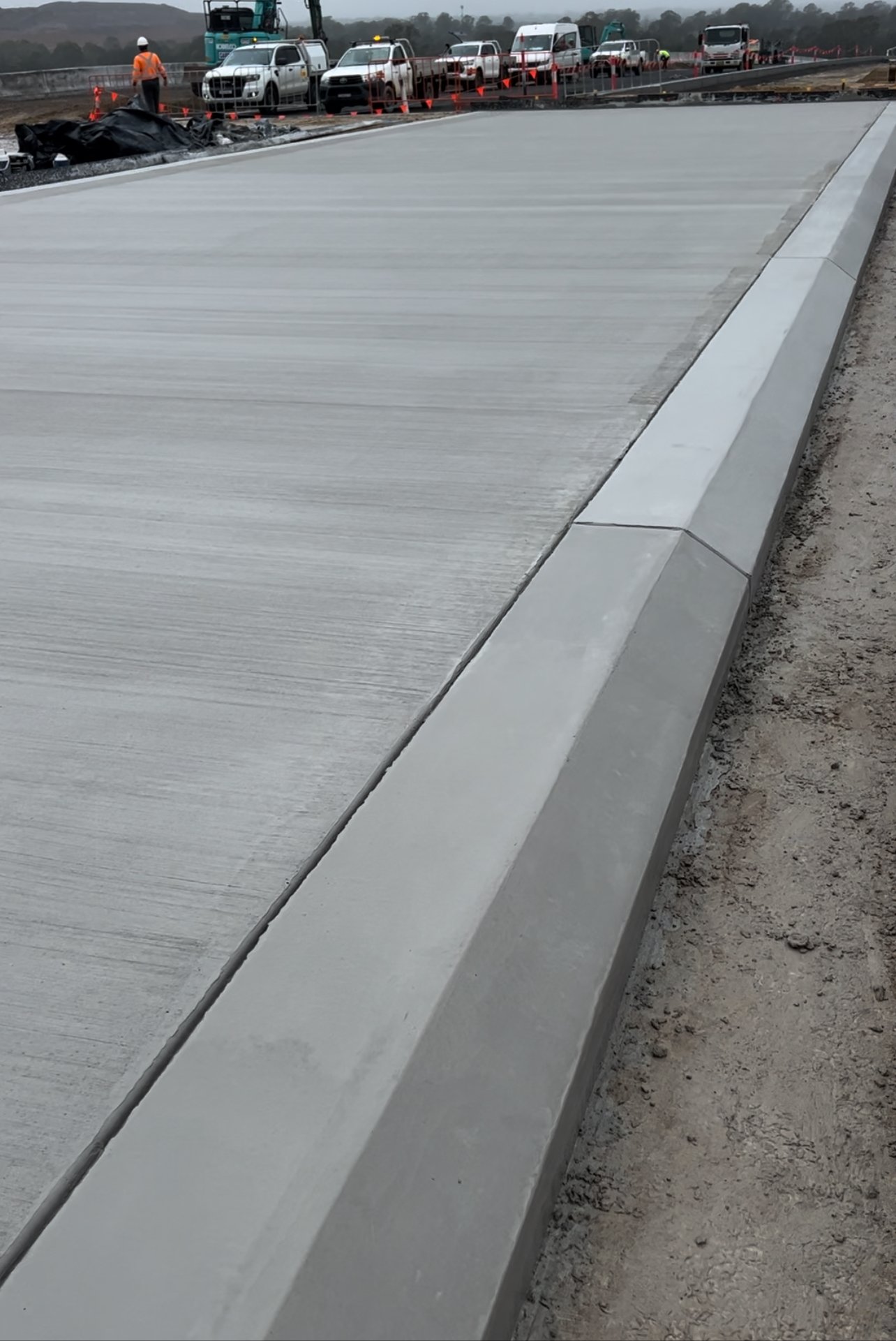 Construction workers and vehicles on a highway under construction, with a newly paved concrete lane and traffic barriers.