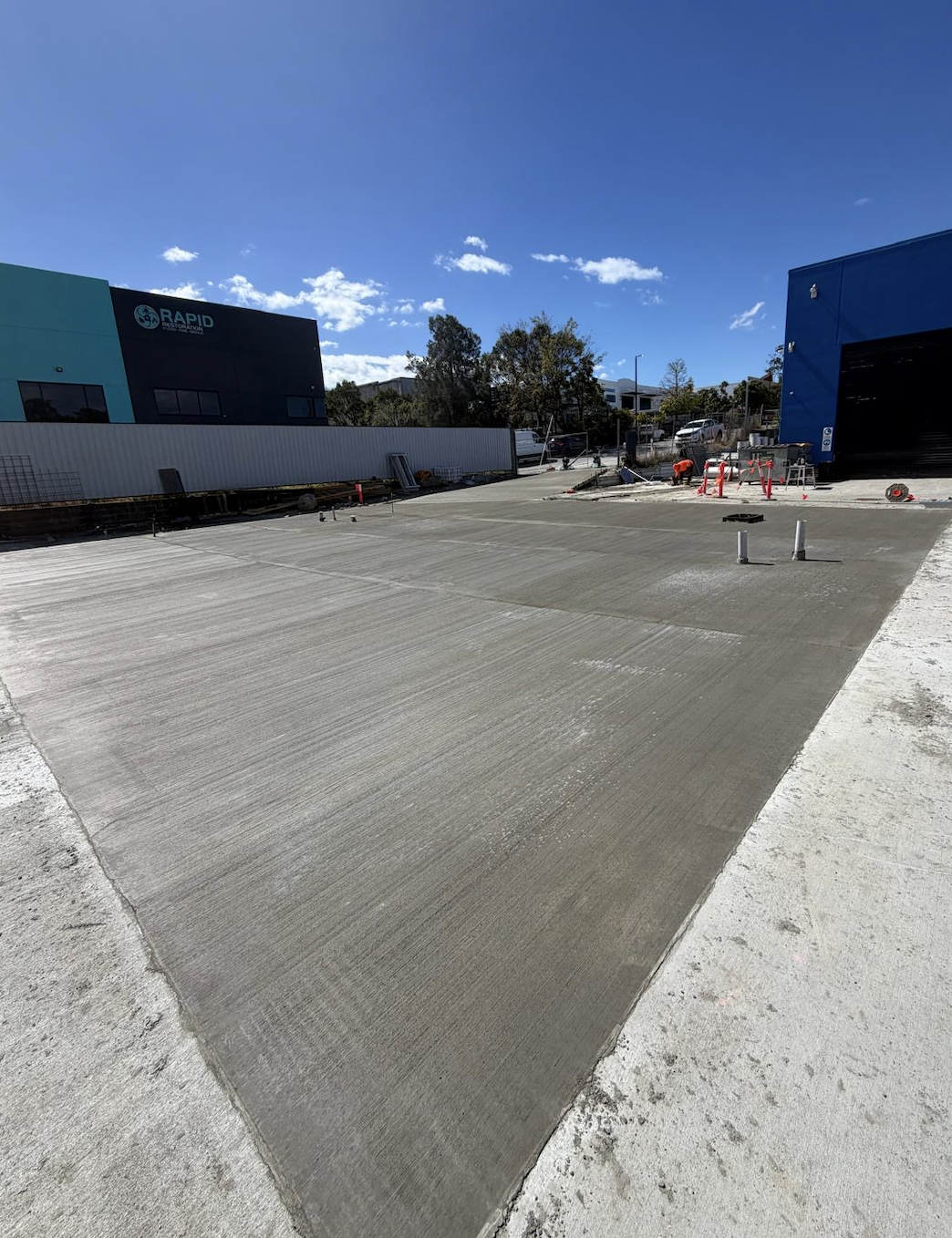 A construction site with fresh concrete on the ground, surrounded by construction materials and orange safety barriers, under a clear blue sky.