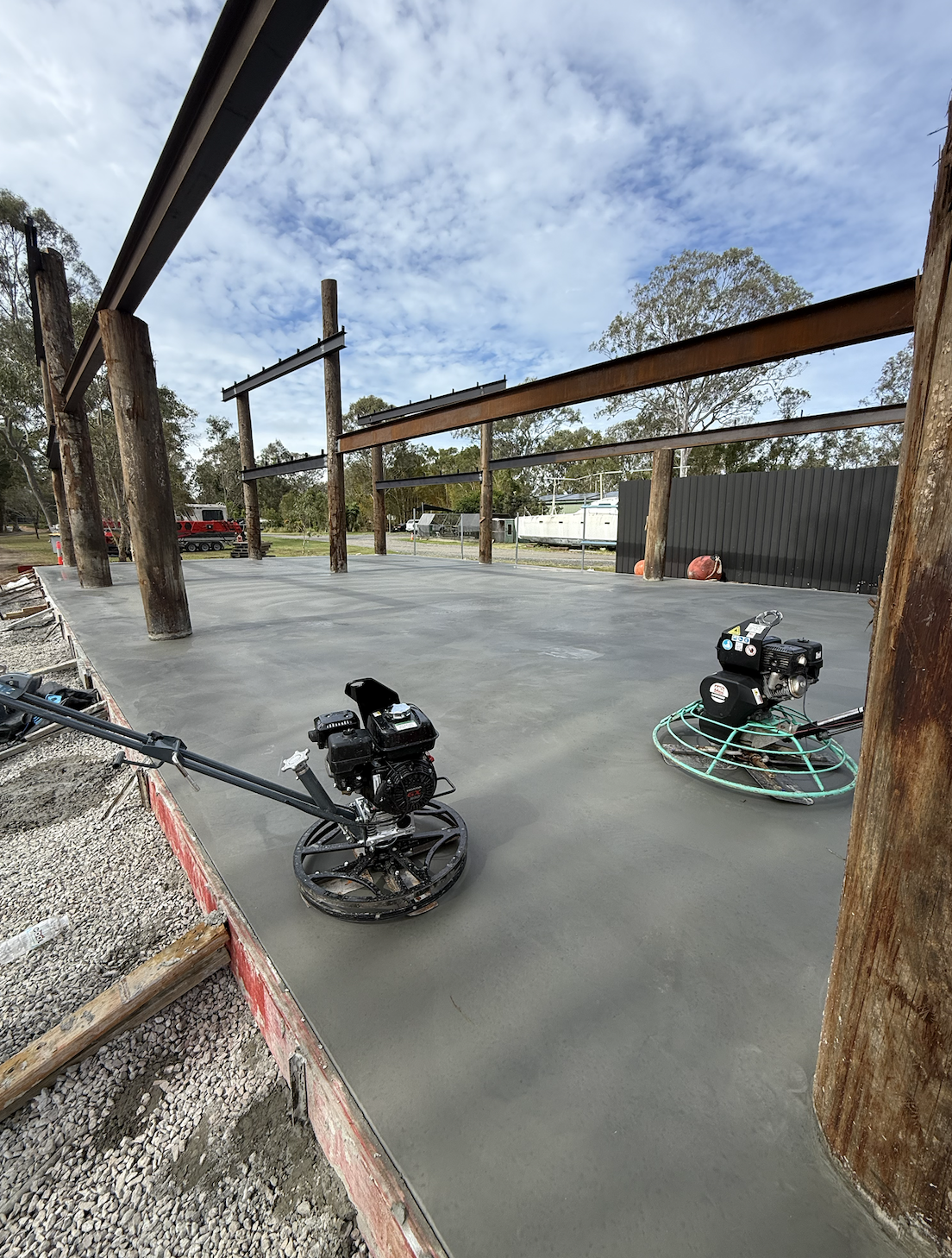 Construction site with freshly poured concrete slab, power trowel machines, wooden support beams, and a partly cloudy sky.