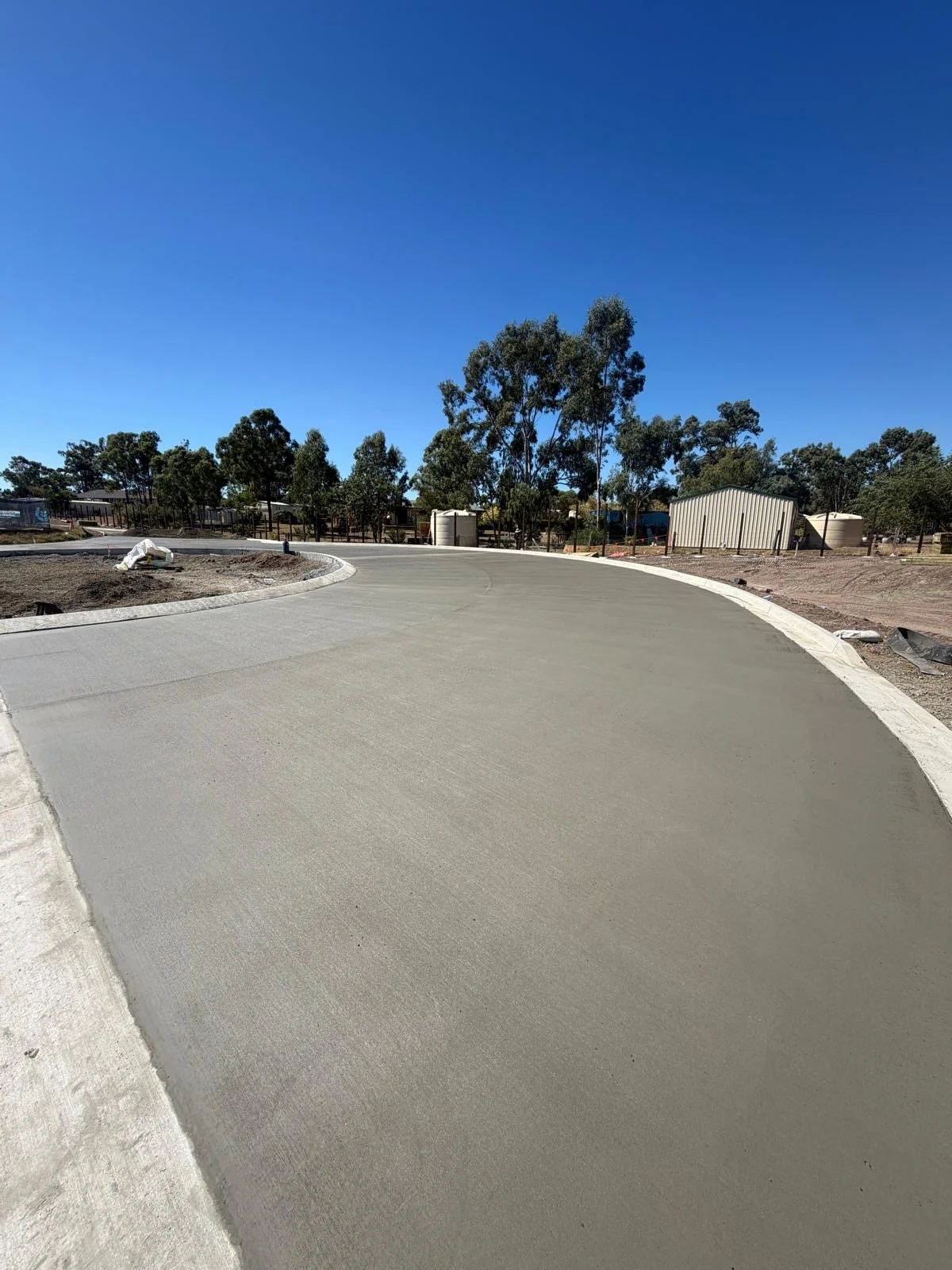 Freshly poured concrete sidewalk with a curved pathway under a clear blue sky, surrounded by sparse trees and construction materials.
