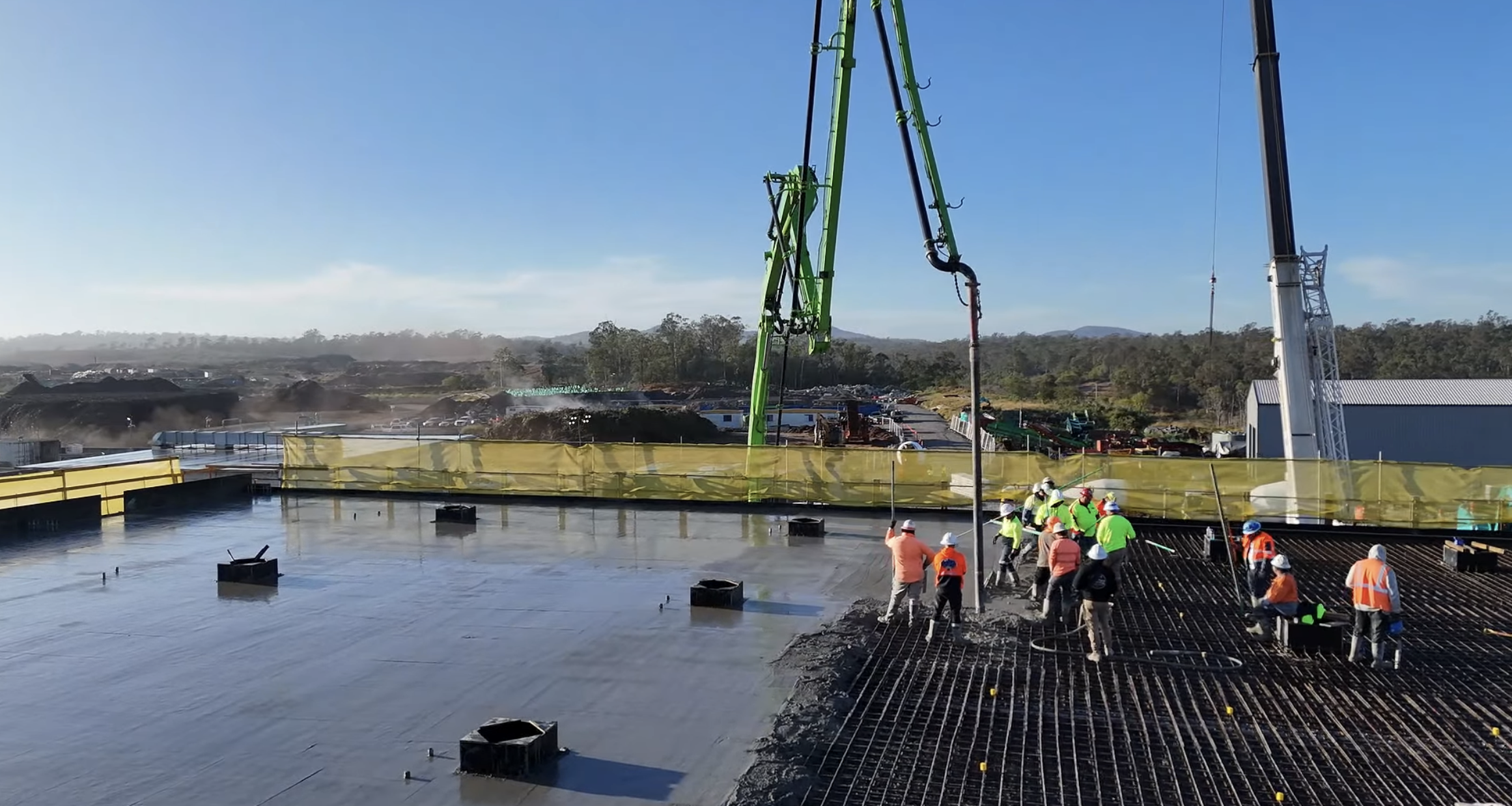 Construction workers in safety gear working on a concrete slab at a construction site with large green and black machinery and a crane in the background, under a clear blue sky.