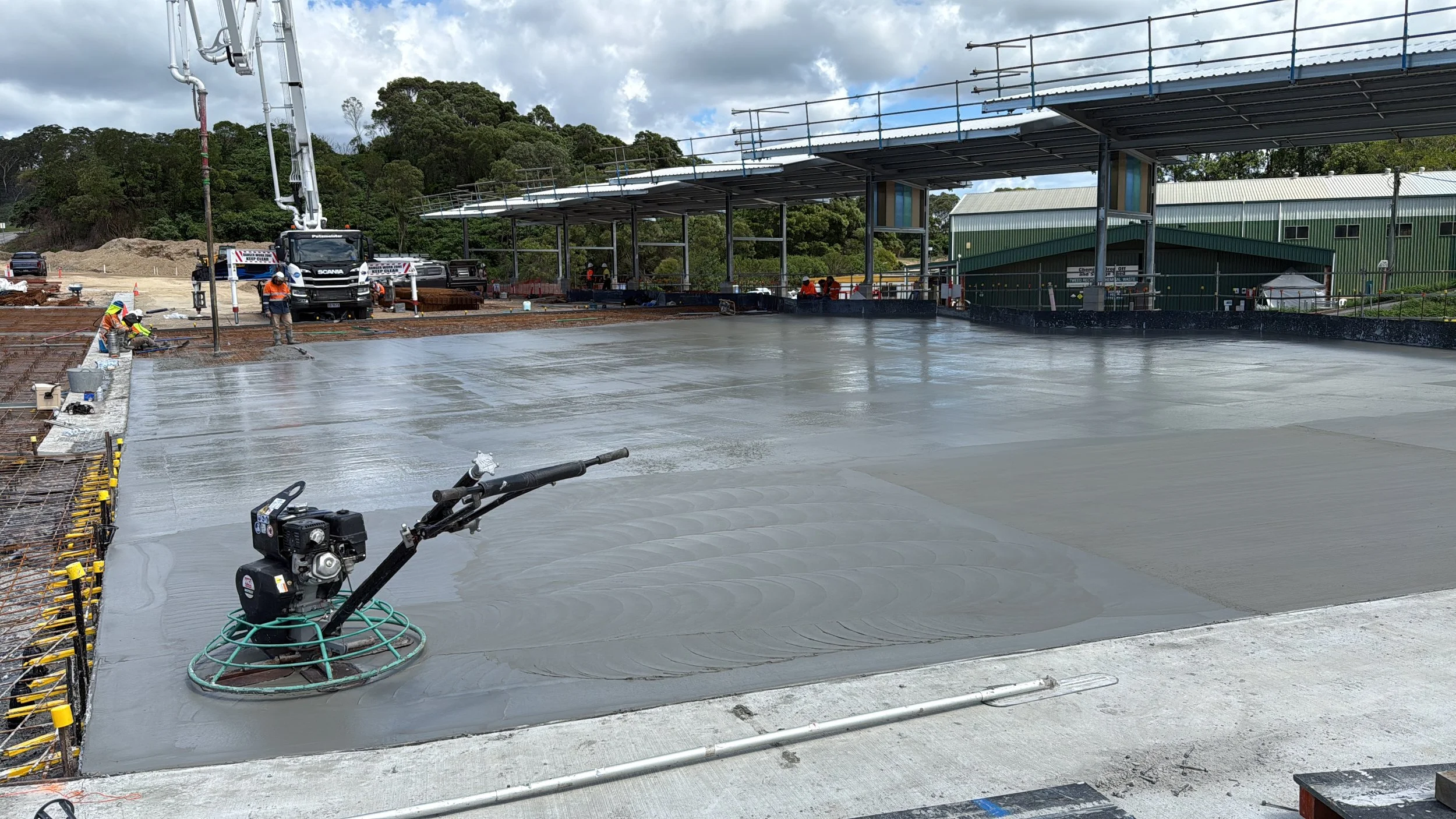 Construction workers pouring and smoothing concrete on a building site, with construction equipment, scaffolding, and a truck in the background.