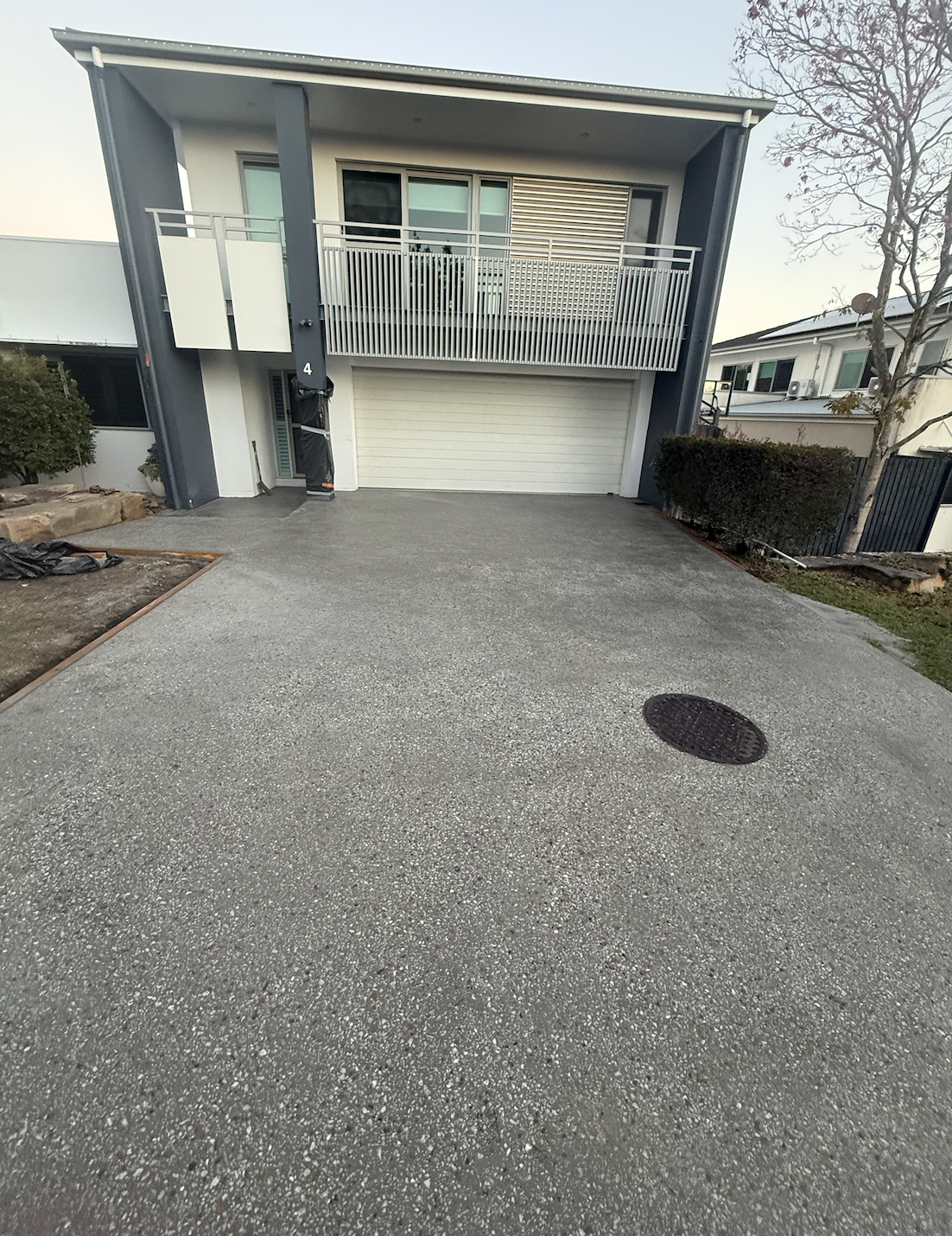 Front of a modern two-story house with a garage and second-floor balcony. The driveway is made of gray gravel, with a drain cover visible in the foreground. There are bushes on the side and a tree without leaves on the right.