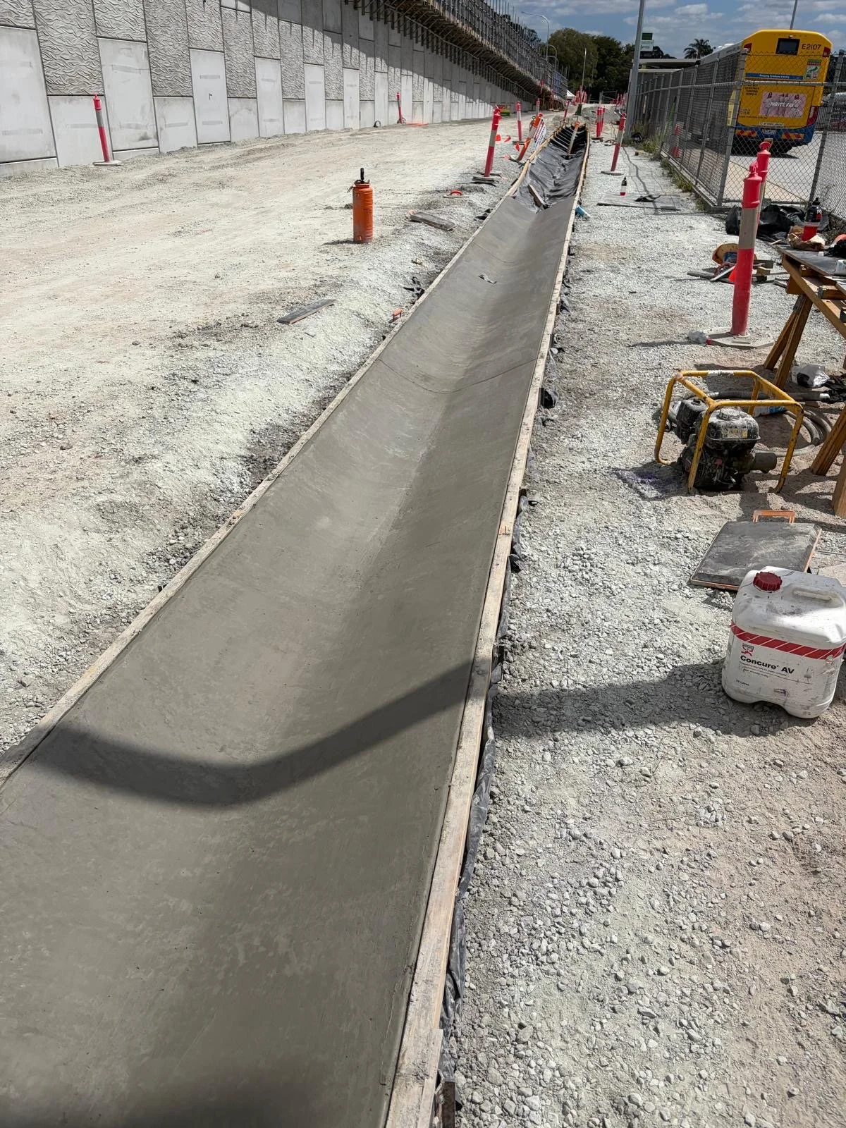 Construction site with fresh concrete sidewalk in progress, surrounded by gravel, construction tools, and safety cones.