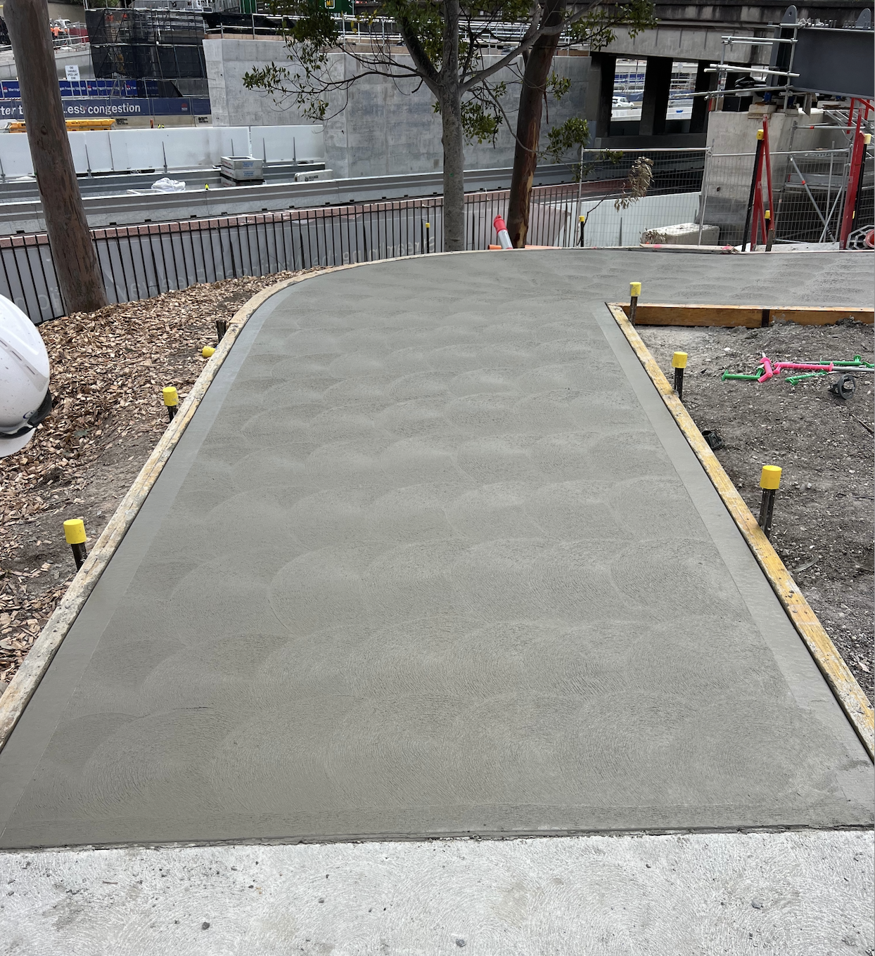 Freshly poured concrete sidewalk with patterned surface in a construction site, surrounded by wooden framing, yellow safety caps on stakes, and construction equipment in background.