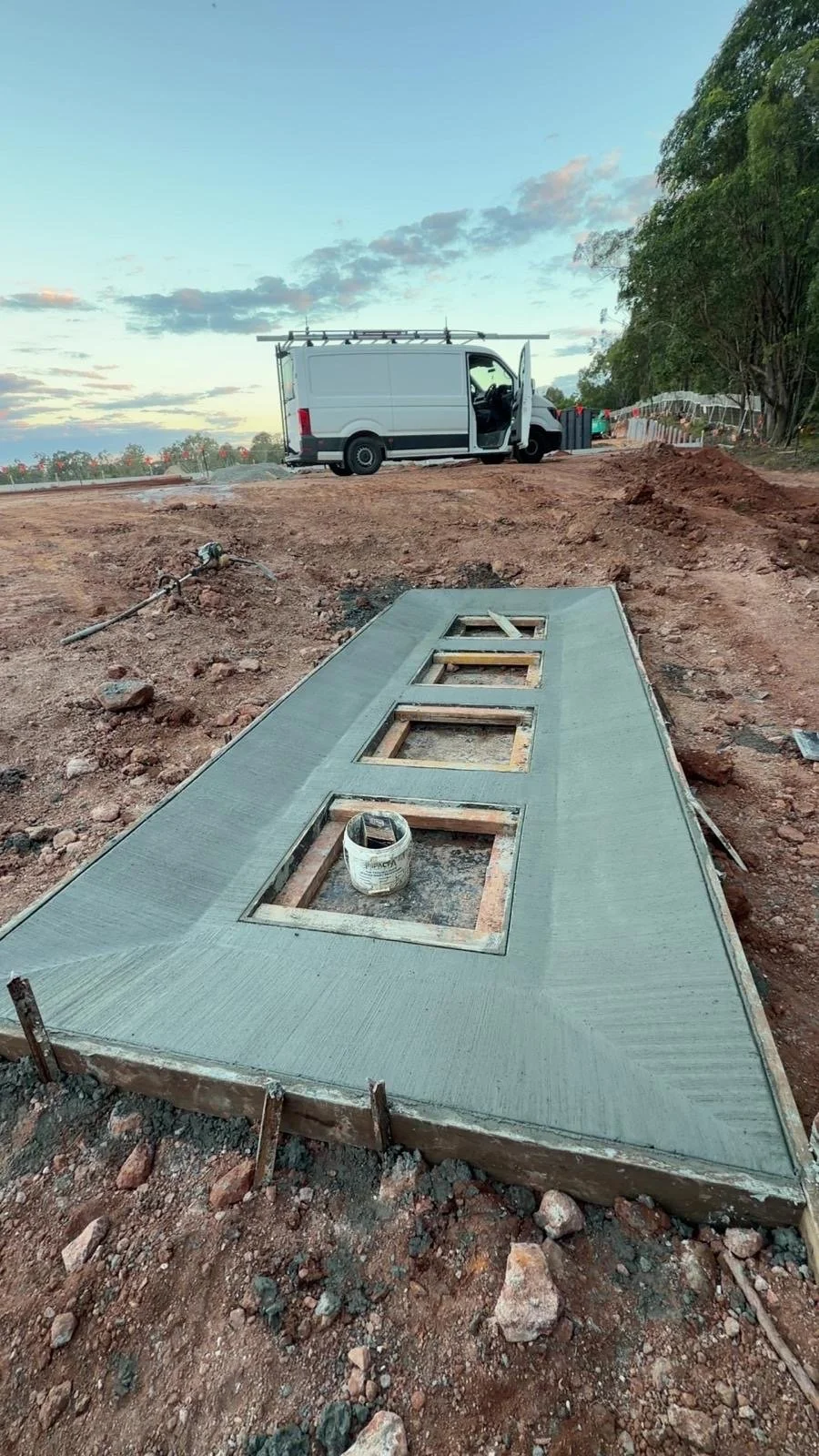 Construction site with a freshly poured concrete slab with four window openings, a white work van parked in the background, and tools on the ground during early evening.
