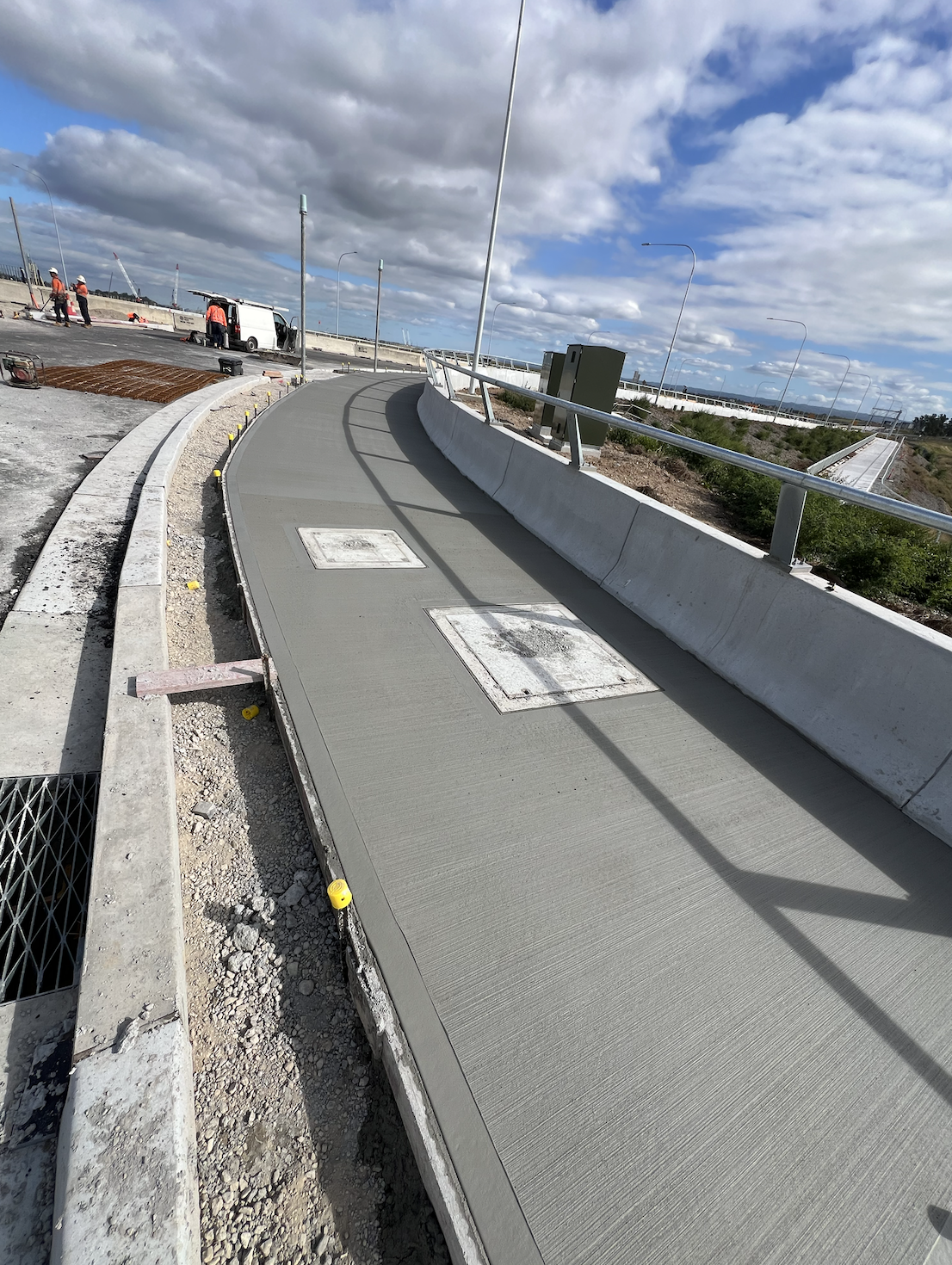 Construction workers paving a sidewalk along a highway under a partly cloudy sky.