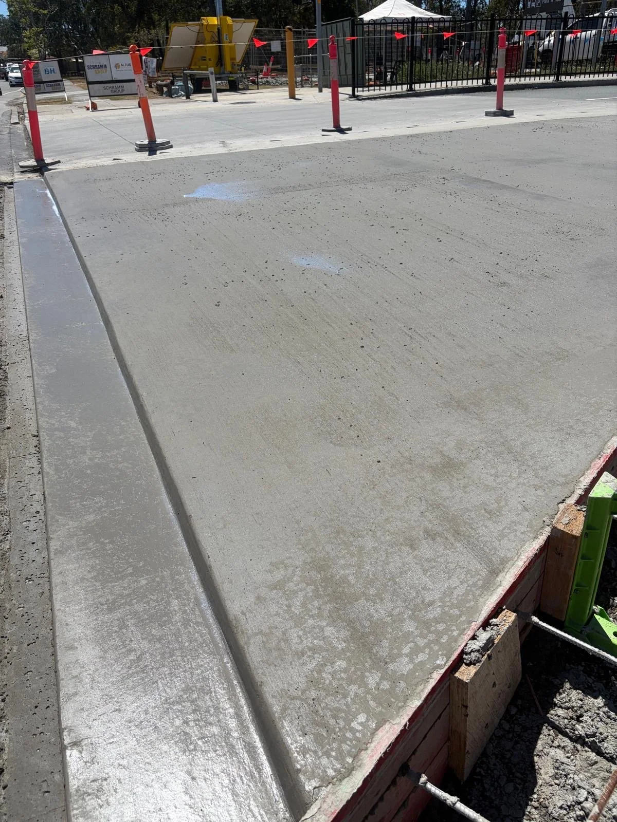 Freshly poured concrete sidewalk with a curb, construction barriers, and equipment visible in the background at a construction site.