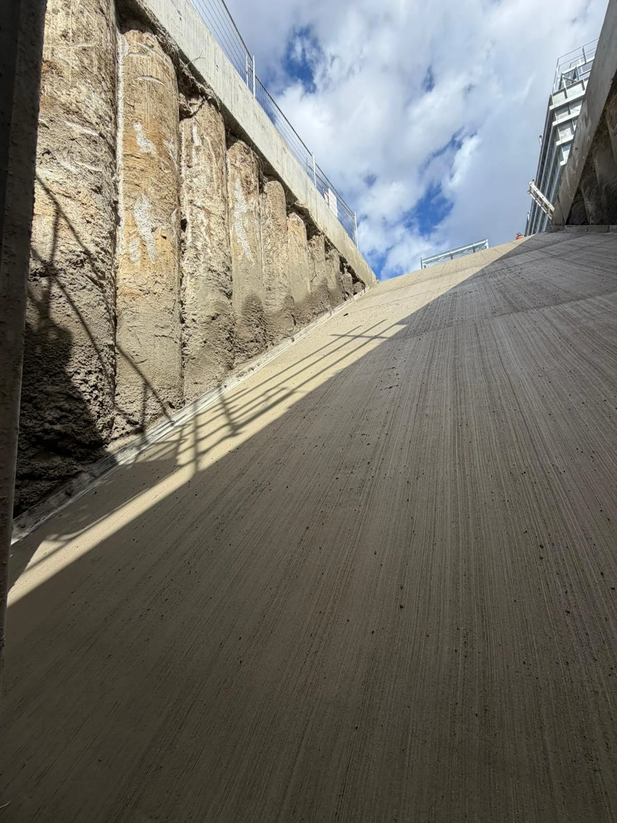 Construction site with a concrete wall on the left and a slope in the foreground, shadows cast by a railing, with a blue sky and clouds overhead.