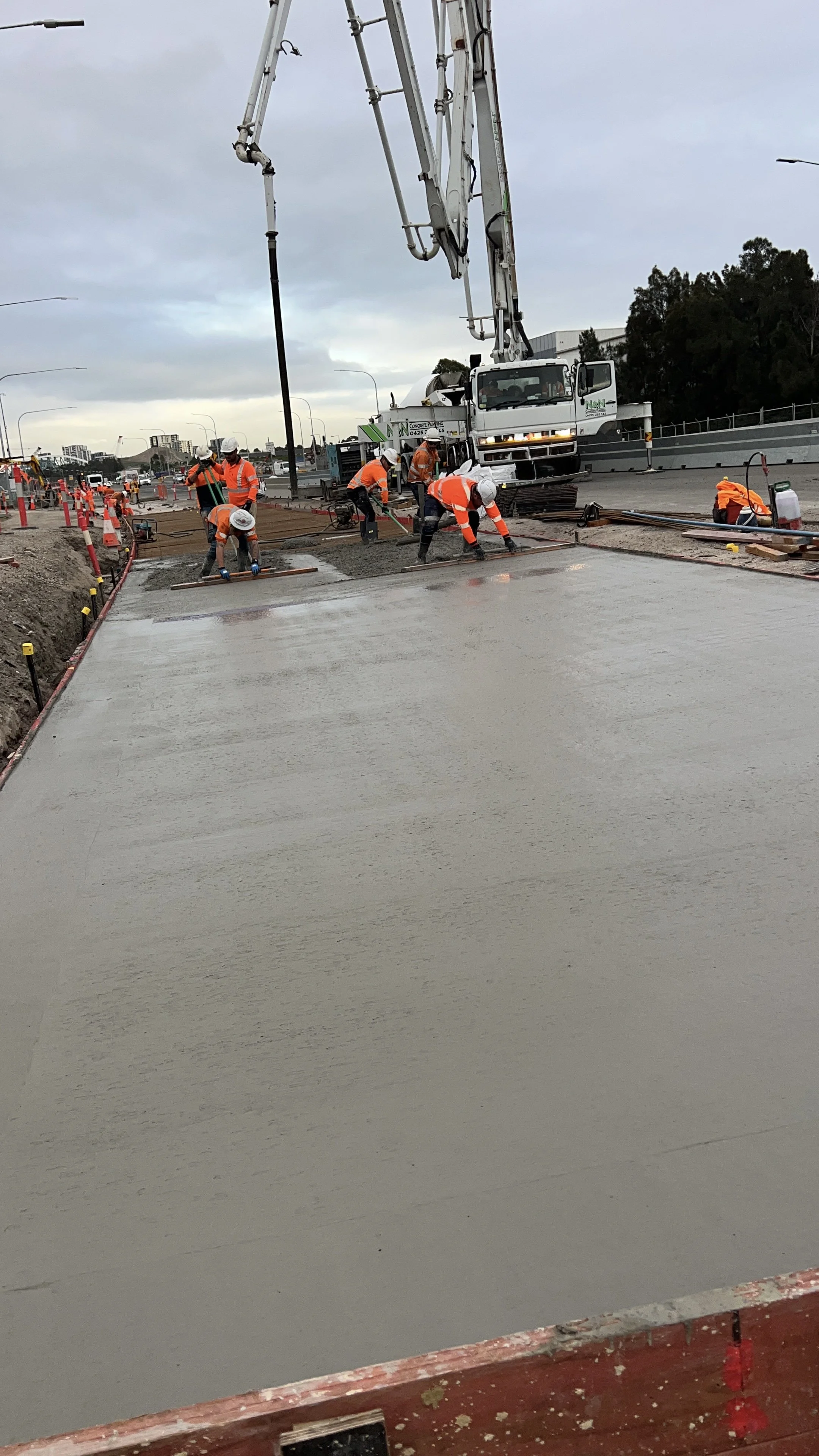 Construction workers in orange safety vests and white helmets pouring and spreading concrete on a road under a cloudy sky, with construction equipment and trucks in the background.