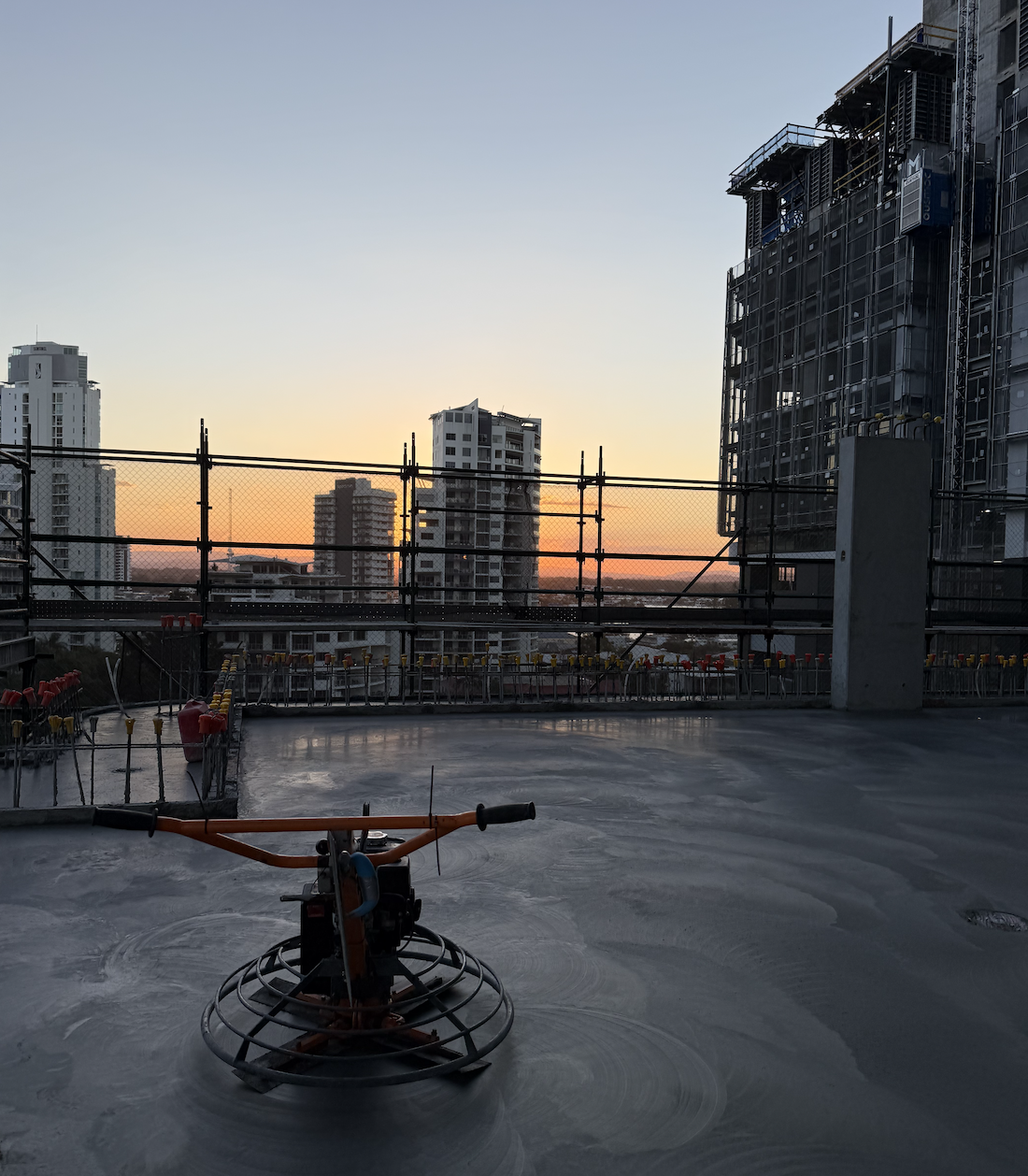 Construction site rooftop at sunset with a concrete finishing machine in the foreground, surrounded by scaffolding, high-rise buildings in the background, and a clear sky with the sun setting on the horizon.