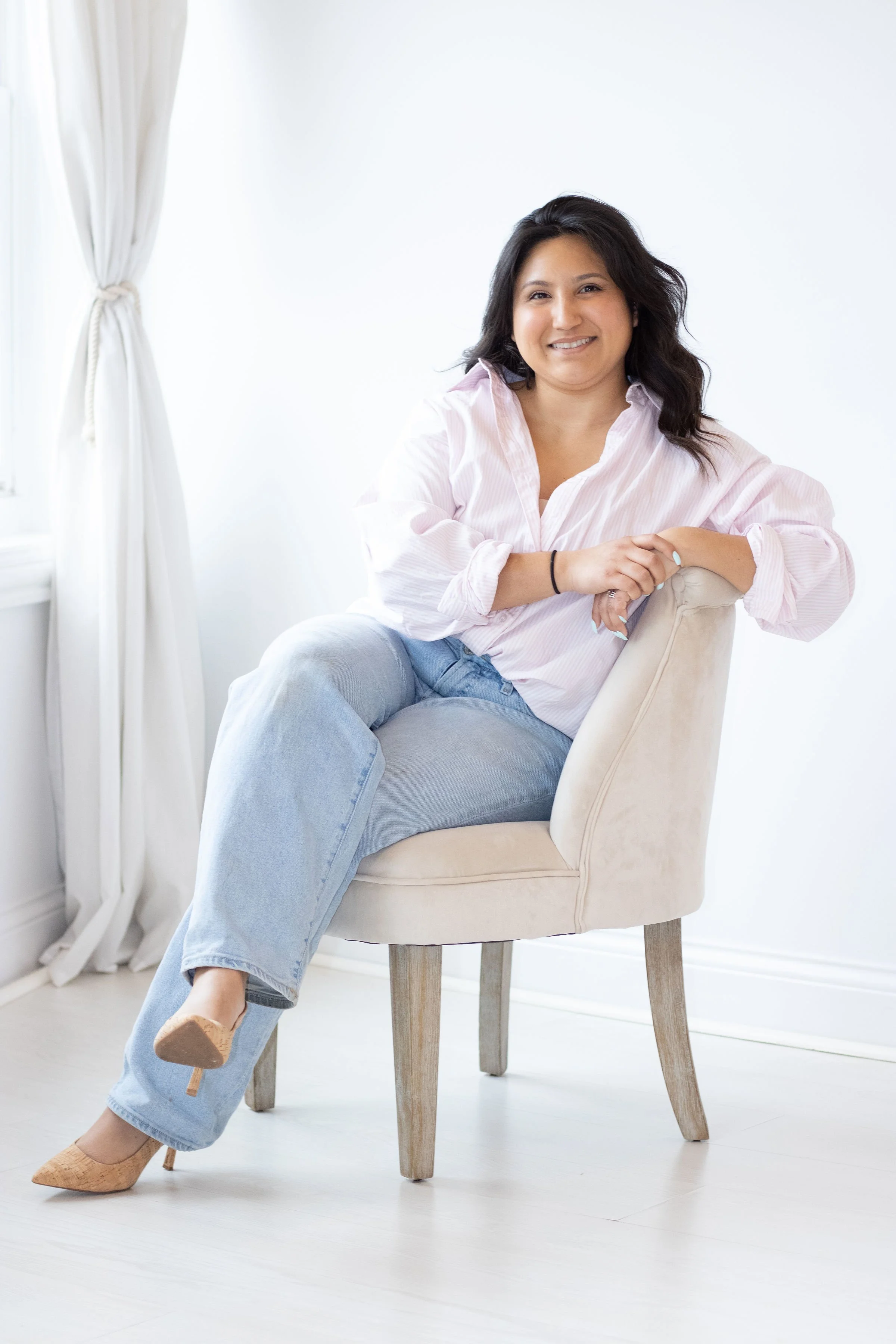 Woman sitting in a beige chair near a window with white curtains, wearing a light pink striped shirt and light blue jeans, smiling with crossed legs.