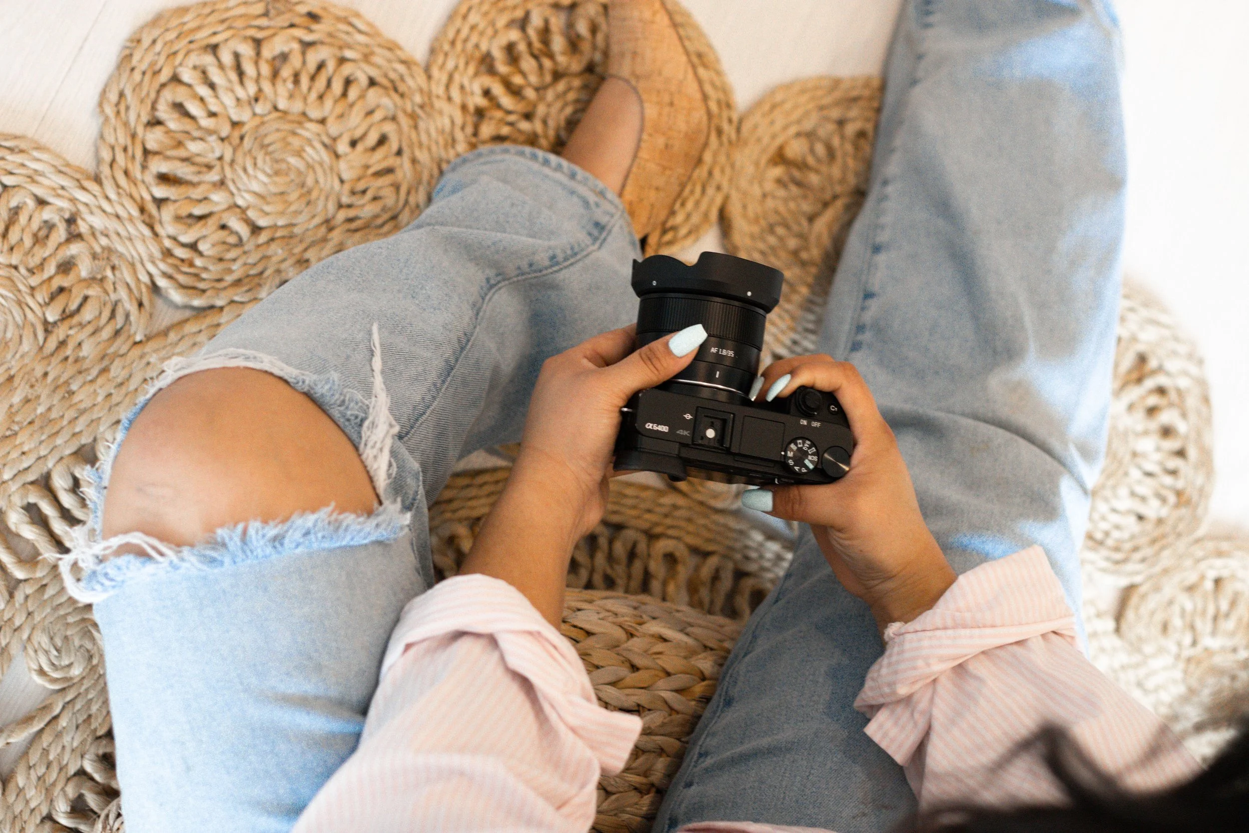 Person sitting on a woven rug, wearing ripped denim shorts and a pink and white striped shirt, holding a black camera with a large lens.