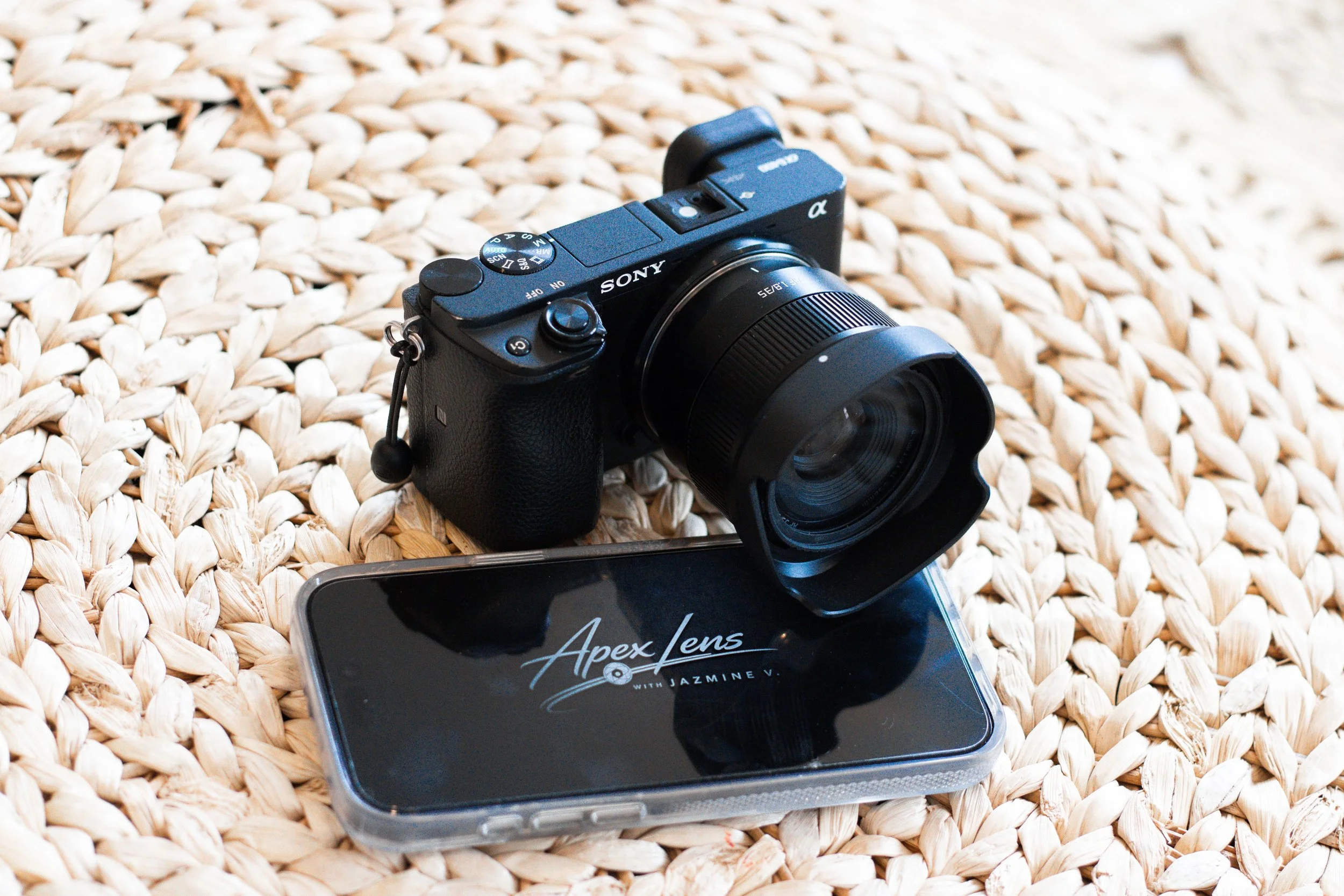 A black Sony camera with a zoom lens resting on a woven surface, with a smartphone underneath and a box labeled 'Apex Lens with Jazmine V.' nearby.