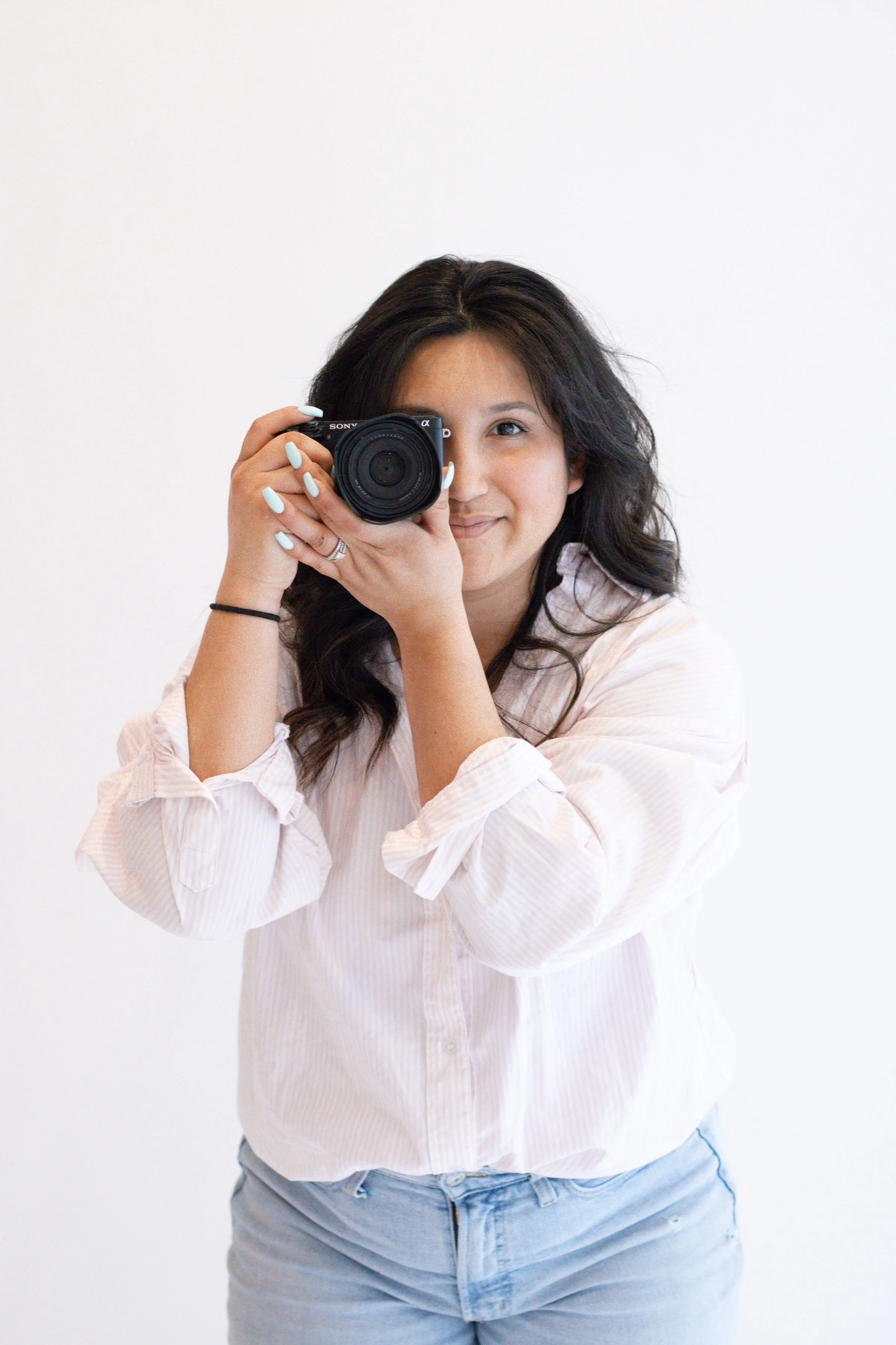 Woman with dark hair smiling, holding a camera up to her face, wearing a light-colored striped shirt and gray jeans, standing against a plain white background.