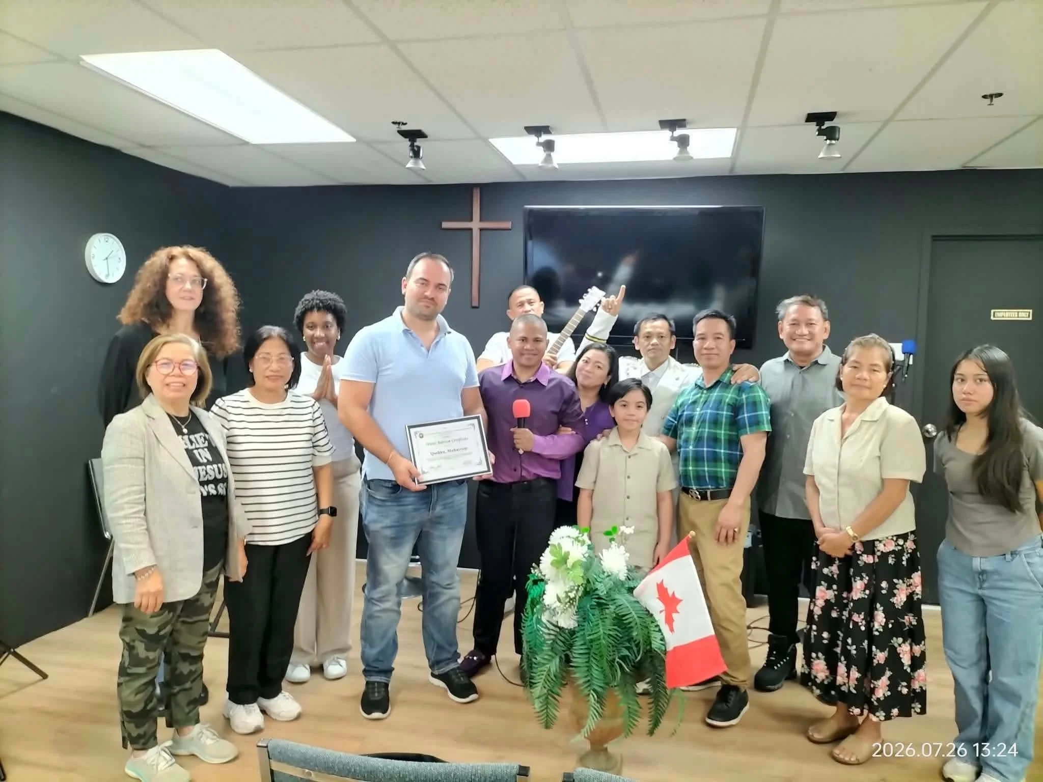 Group of diverse people gathered in a room with a cross on the wall, some holding certificates and others smiling or giving a thumbs-up, with a large TV screen behind them and a flower arrangement with a Canadian flag in the foreground.