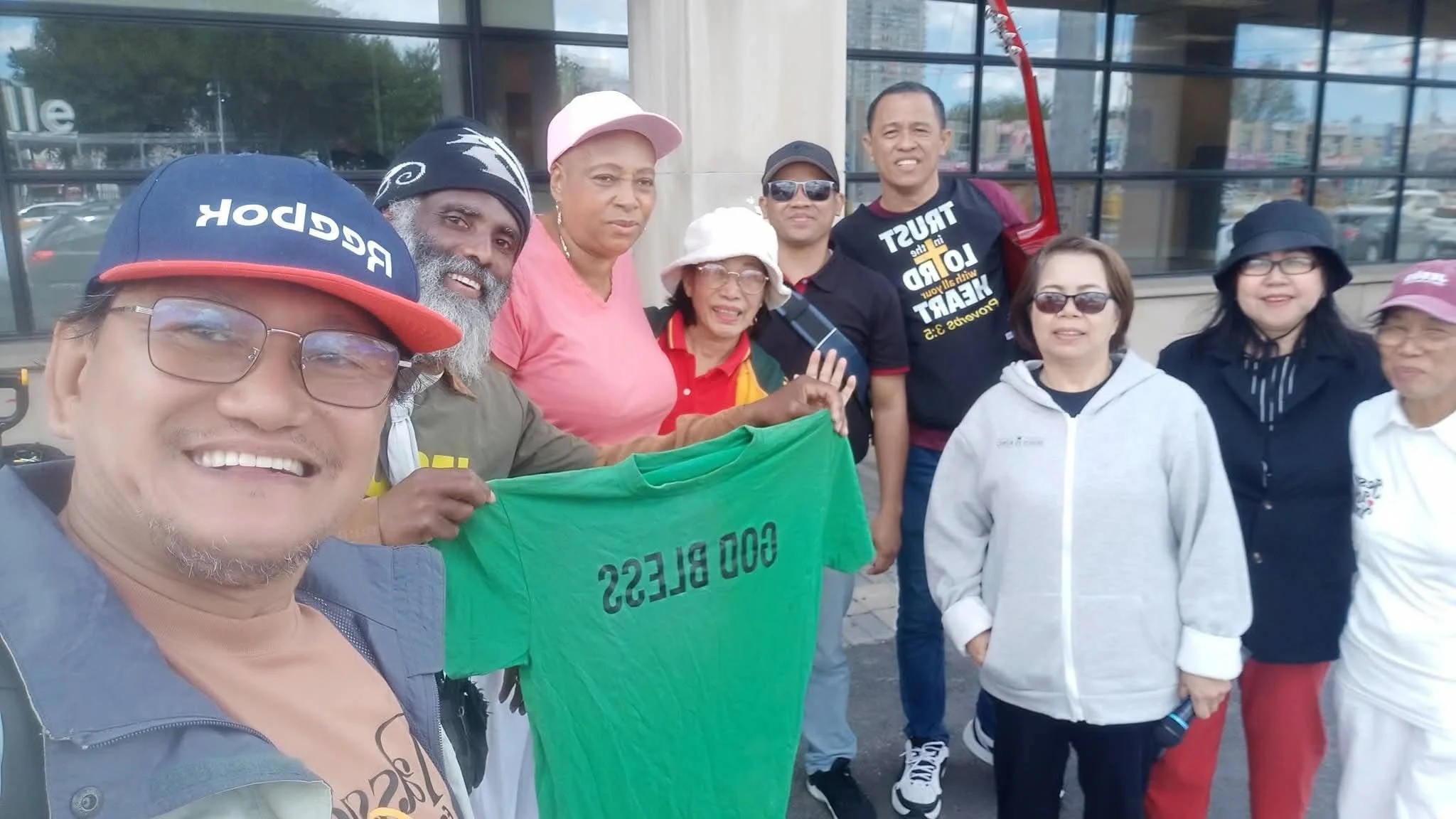 Group of people standing together outside a building, holding a green banner that reads 'GOO BLESS'.