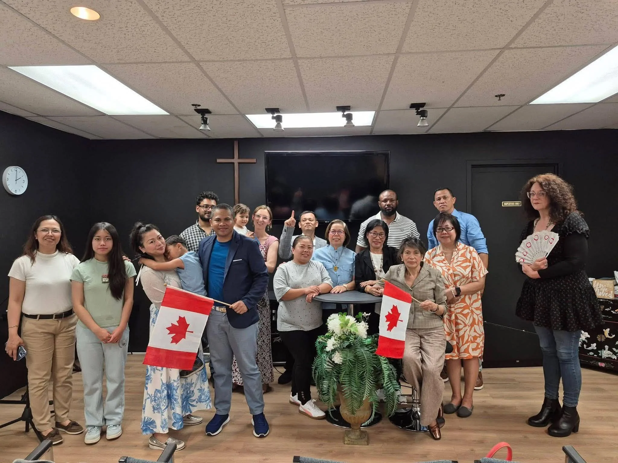 A diverse group of people gathered in a room with a black wall and a clock, holding Canadian flags, with a cross on the black wall and a large TV behind them. The group includes men, women, and children of different ages and ethnicities, and a woman on the far right holds a fan.