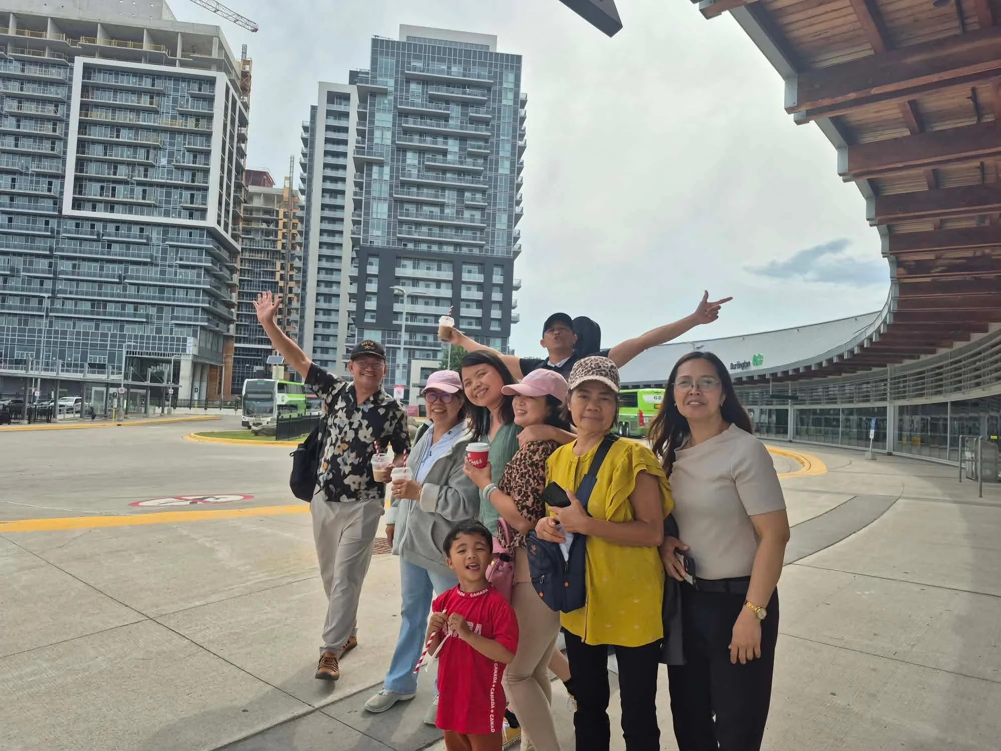 A group of people, including children and adults, standing outdoors at a transportation station with high-rise buildings in the background. Some of the people are smiling and making gestures, holding drinks. The sky is cloudy.