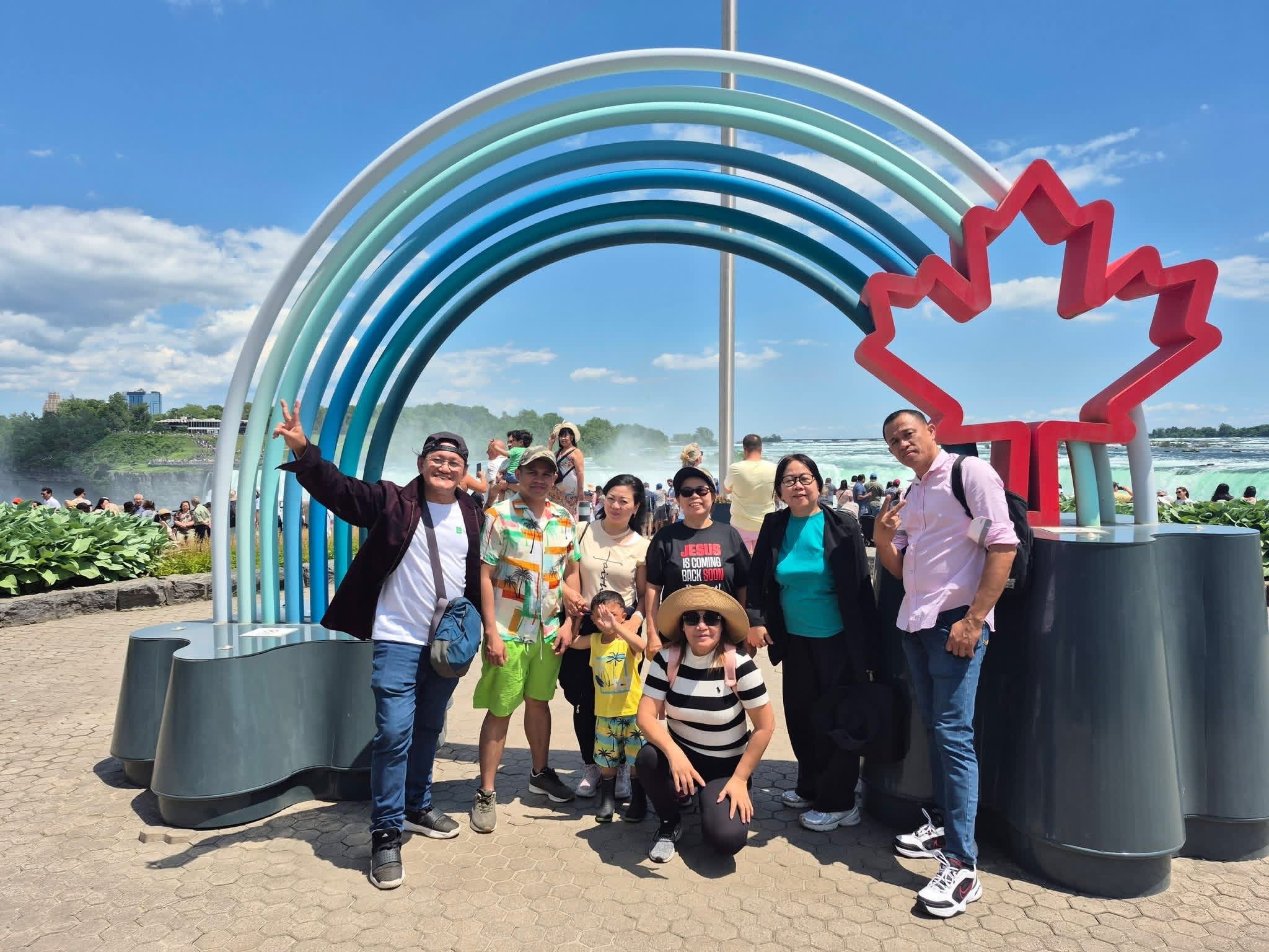 A diverse group of people posing for a photo at a tourist attraction with a large rainbow-shaped sculpture and a red maple leaf outline behind them, under a blue sky with scattered clouds.