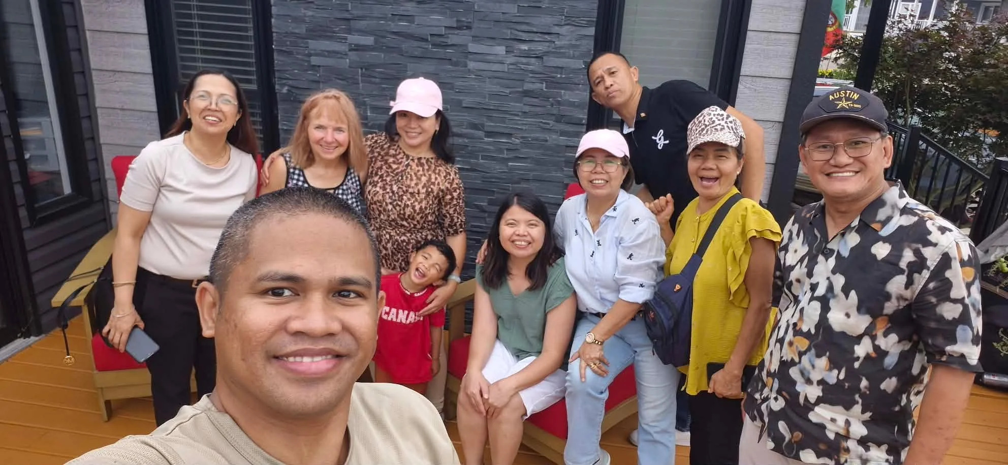 Group of people taking a selfie at an outdoor gathering on a patio with a gray stone wall background. Everyone is smiling and dressed casually.