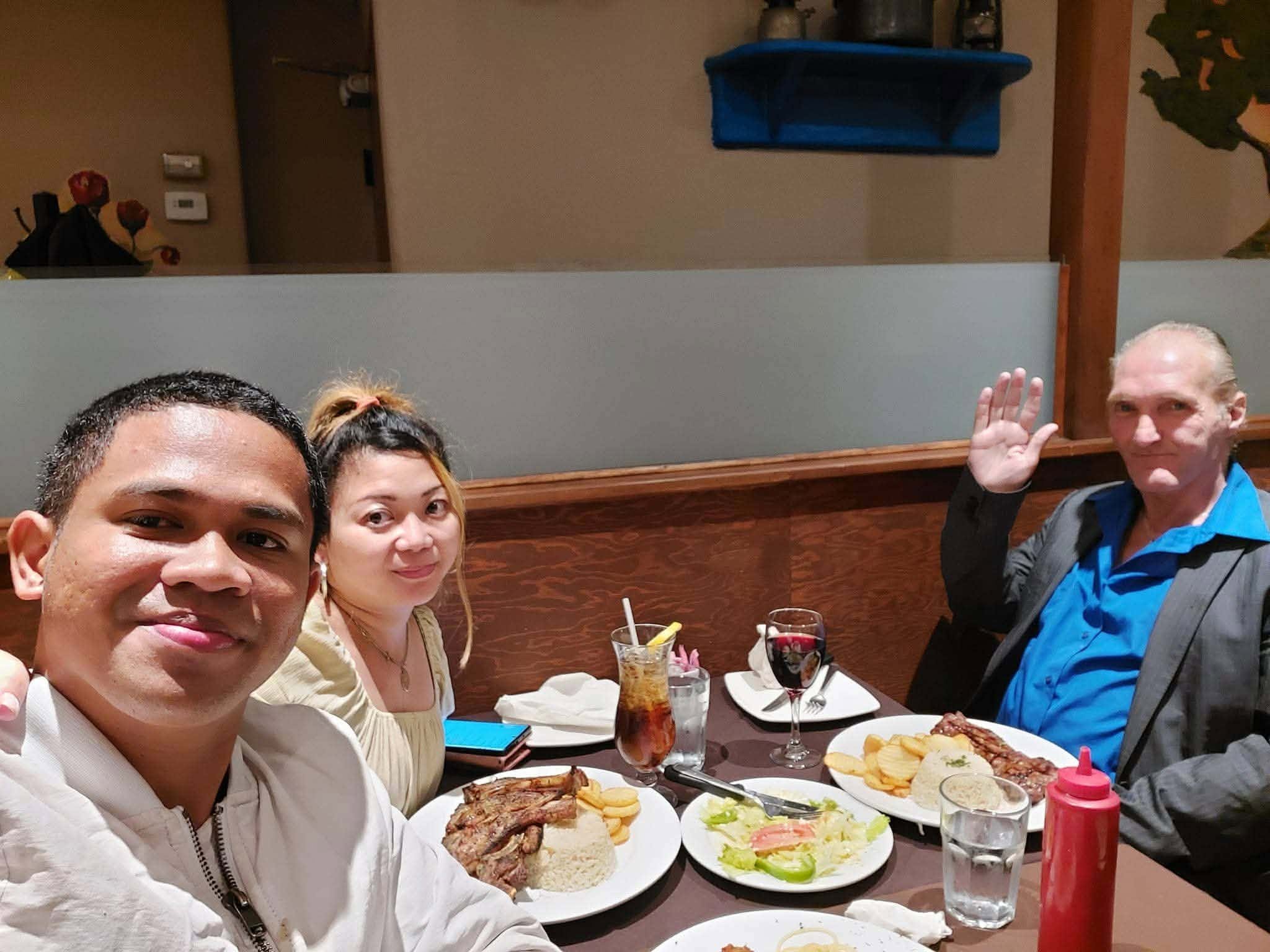 Group of people dining at a restaurant table filled with plates of food, drinks, and condiments, with three people visible in the foreground smiling and posing for the photo.
