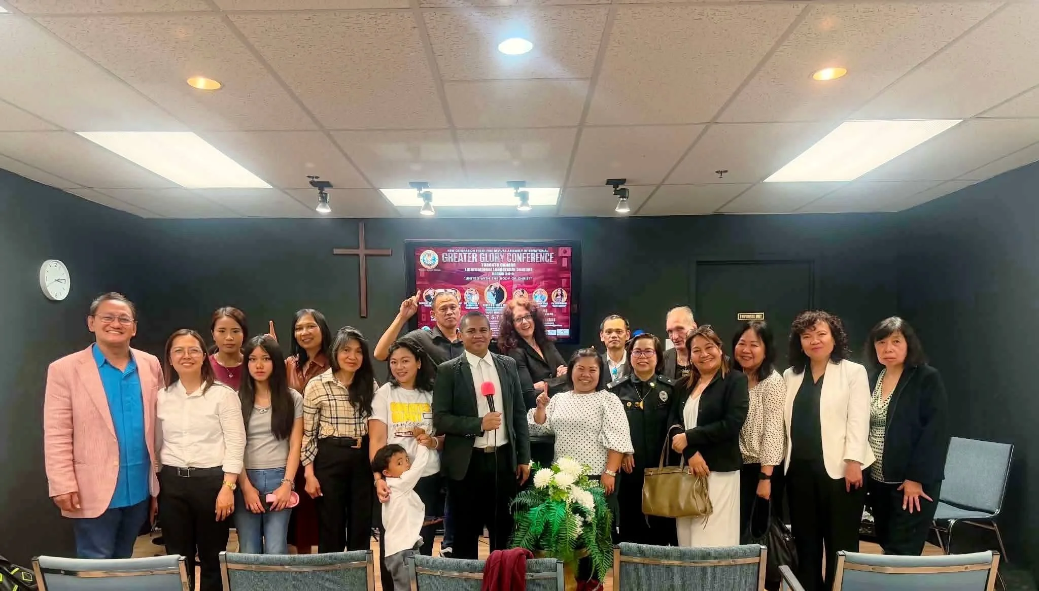 A group of diverse people standing together in a room, smiling for a photo, with a cross on the wall and a TV screen displaying 'Greater Glory Conference' in the background.