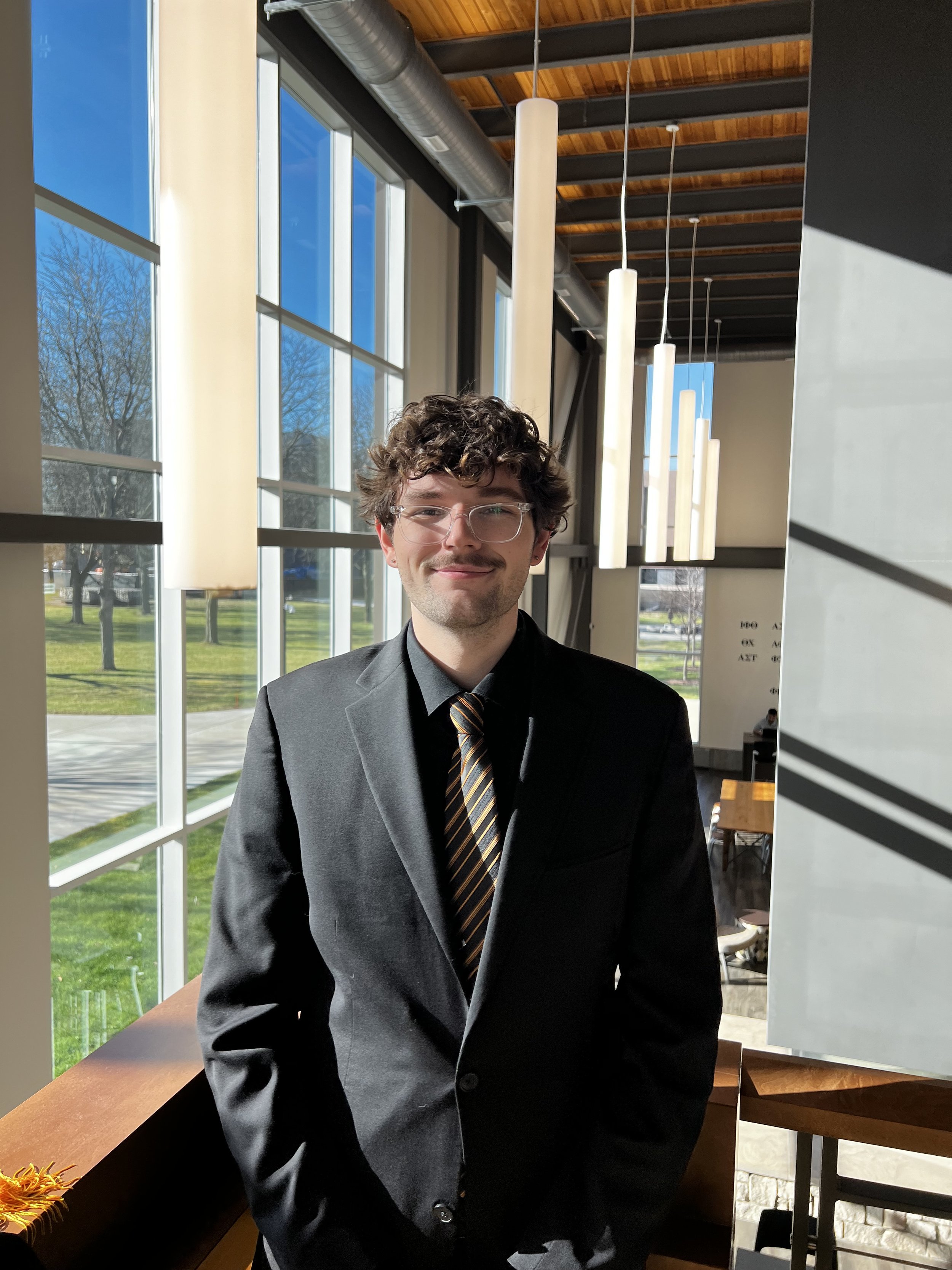 A young man with curly brown hair, glasses, and a light beard is smiling while wearing a black suit, shirt, and striped tie. He is standing indoors near large windows, with sunlight illuminating his face and a view of trees and a grassy area outside.