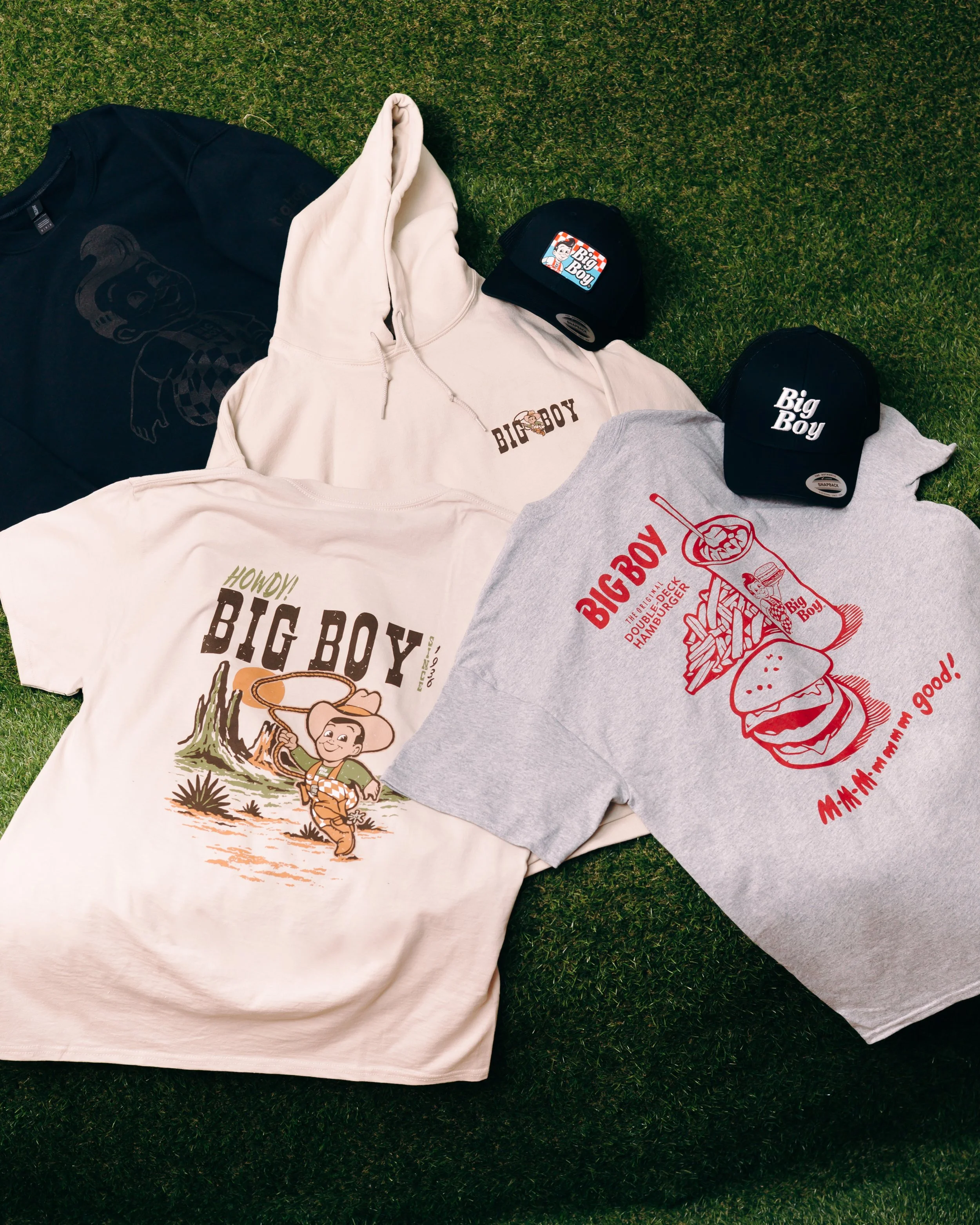 Various Big Boy themed clothing and accessories laid out on artificial grass, including black and gray caps and a hoodie with Big Boy characters and logos.